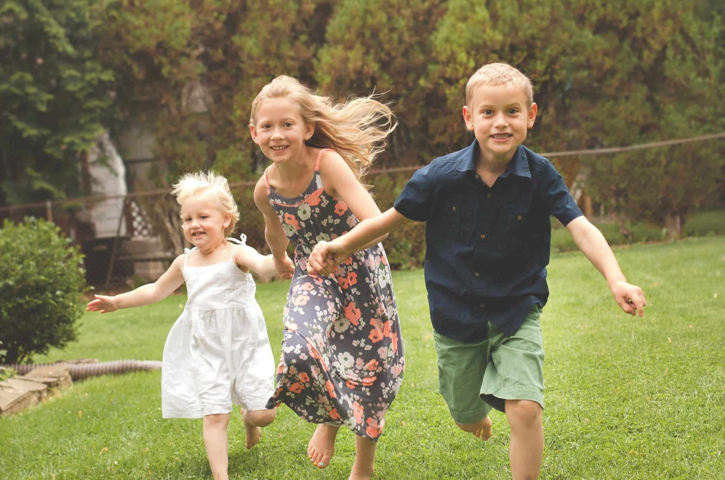 Three children, two girls and one boy, are running and playing outdoors on a grassy yard, holding hands and smiling.
