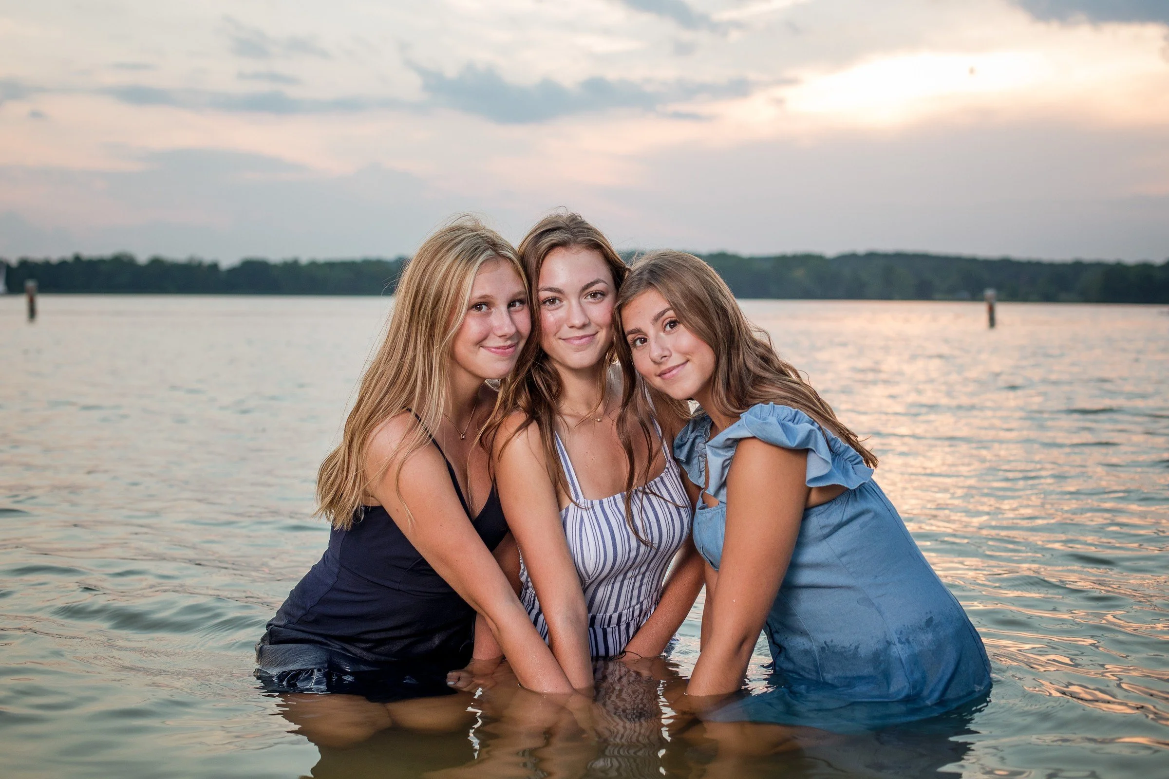 Three young women standing in a body of water during sunset, smiling and close together.