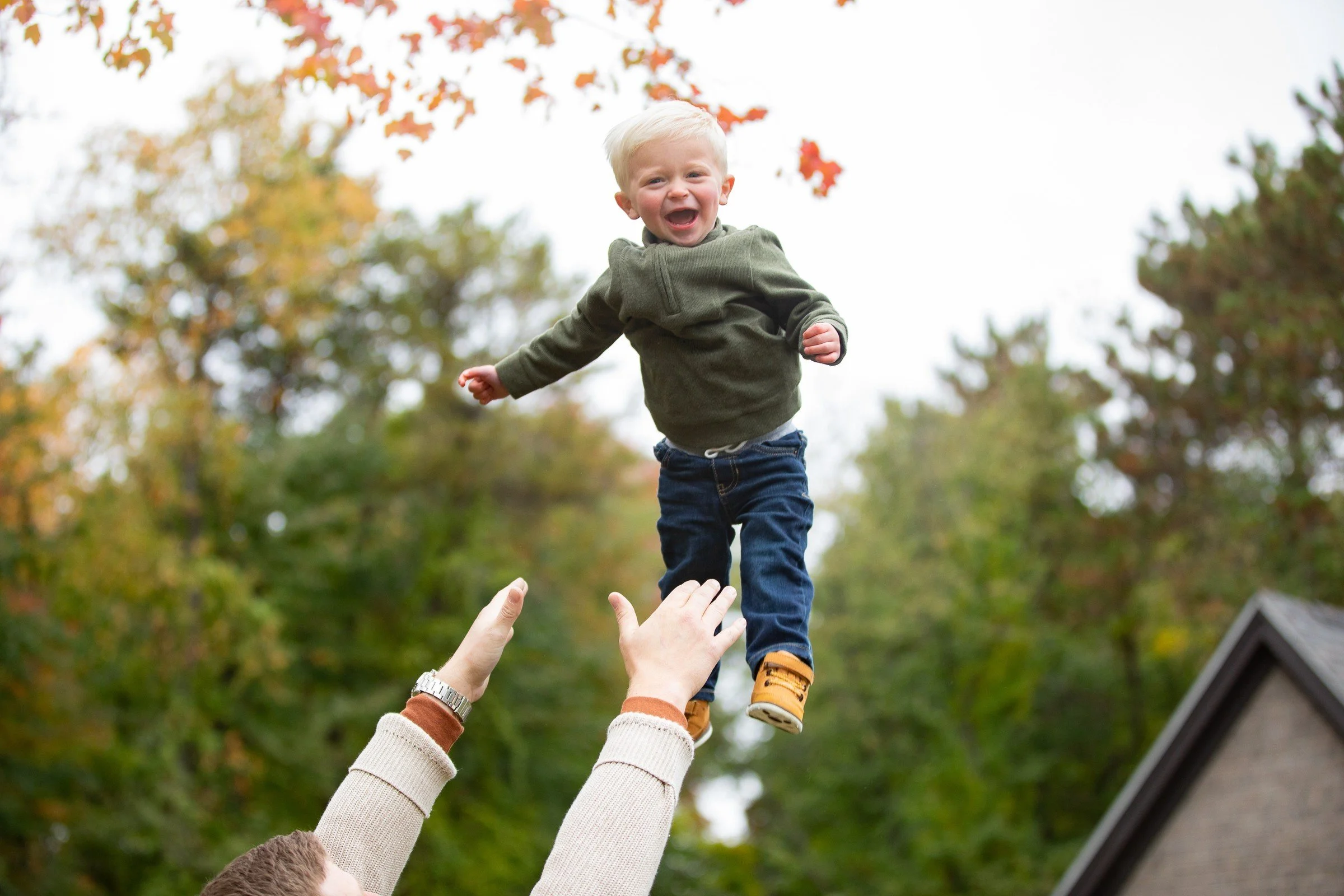 A young blonde boy laughing and joyfully falling from an adult's hands outside during autumn, with trees with fall foliage in the background.