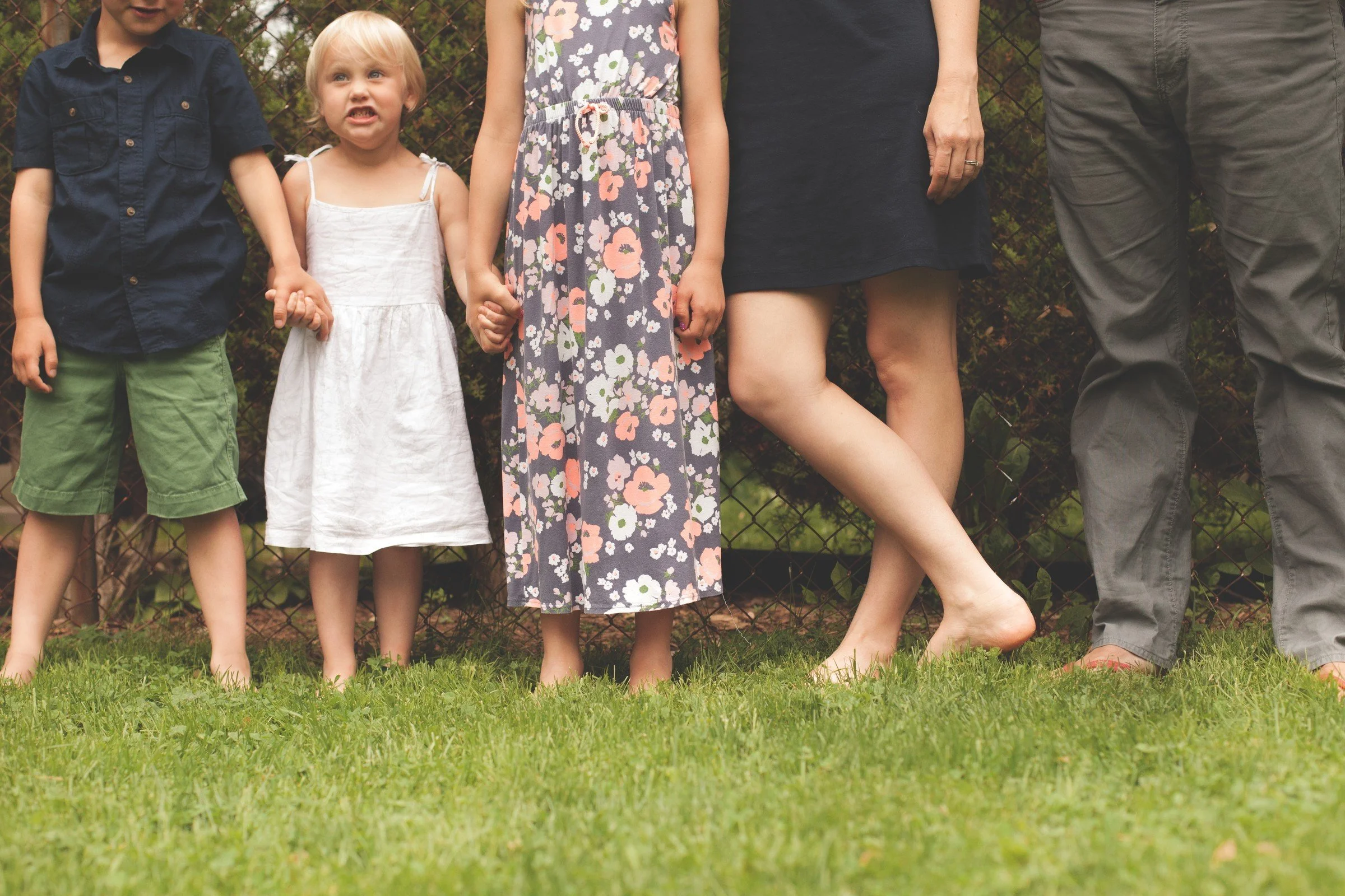 Family standing outdoors near a chain-link fence, with the focus on their lower bodies and legs, on grass.