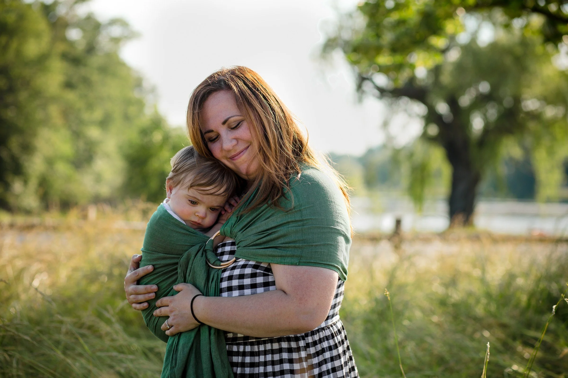 Woman holding a young child in a baby sling outdoors in a grassy area with trees in the background.