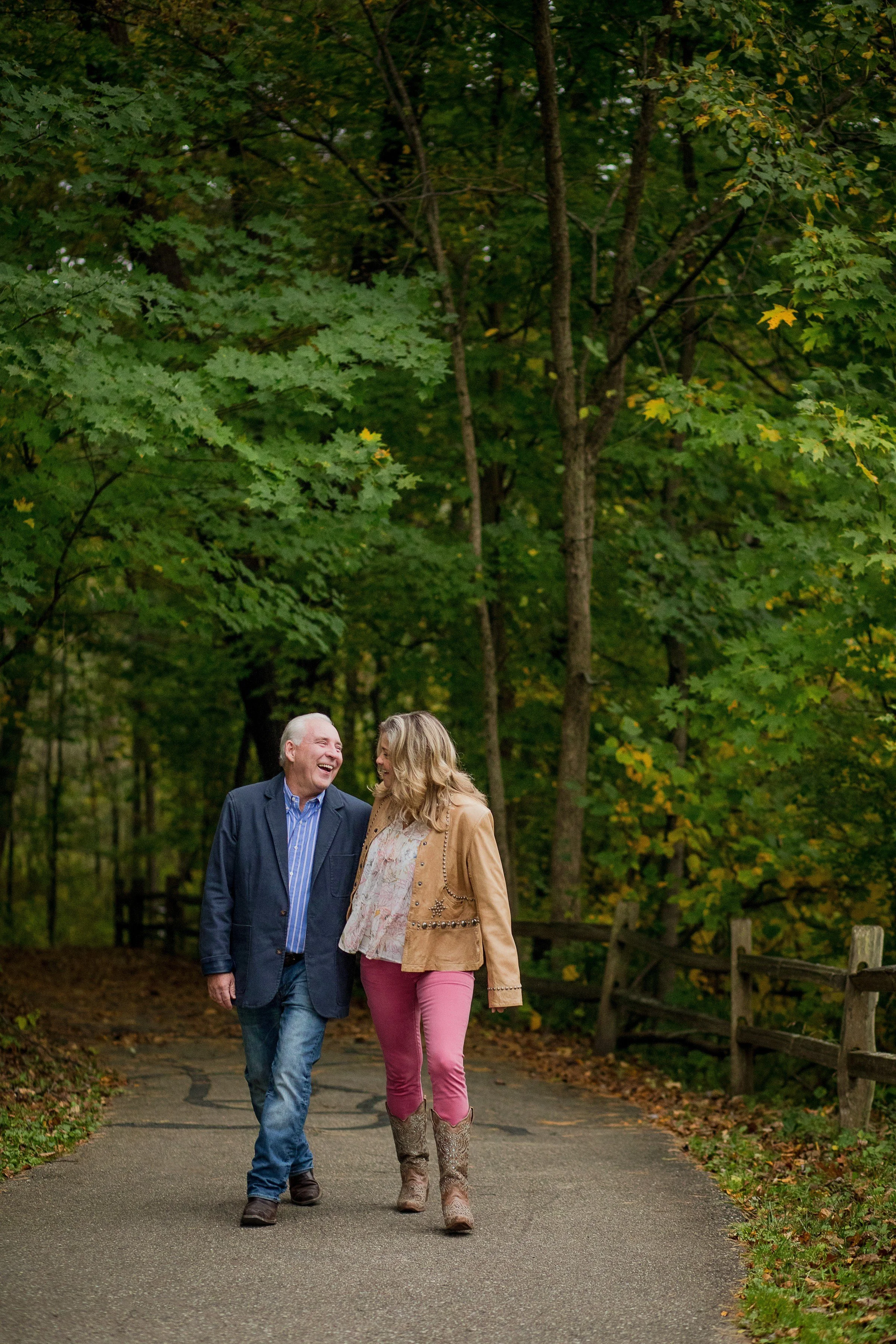A man and woman walk and laugh together on a paved trail through a lush green forest with tall trees.