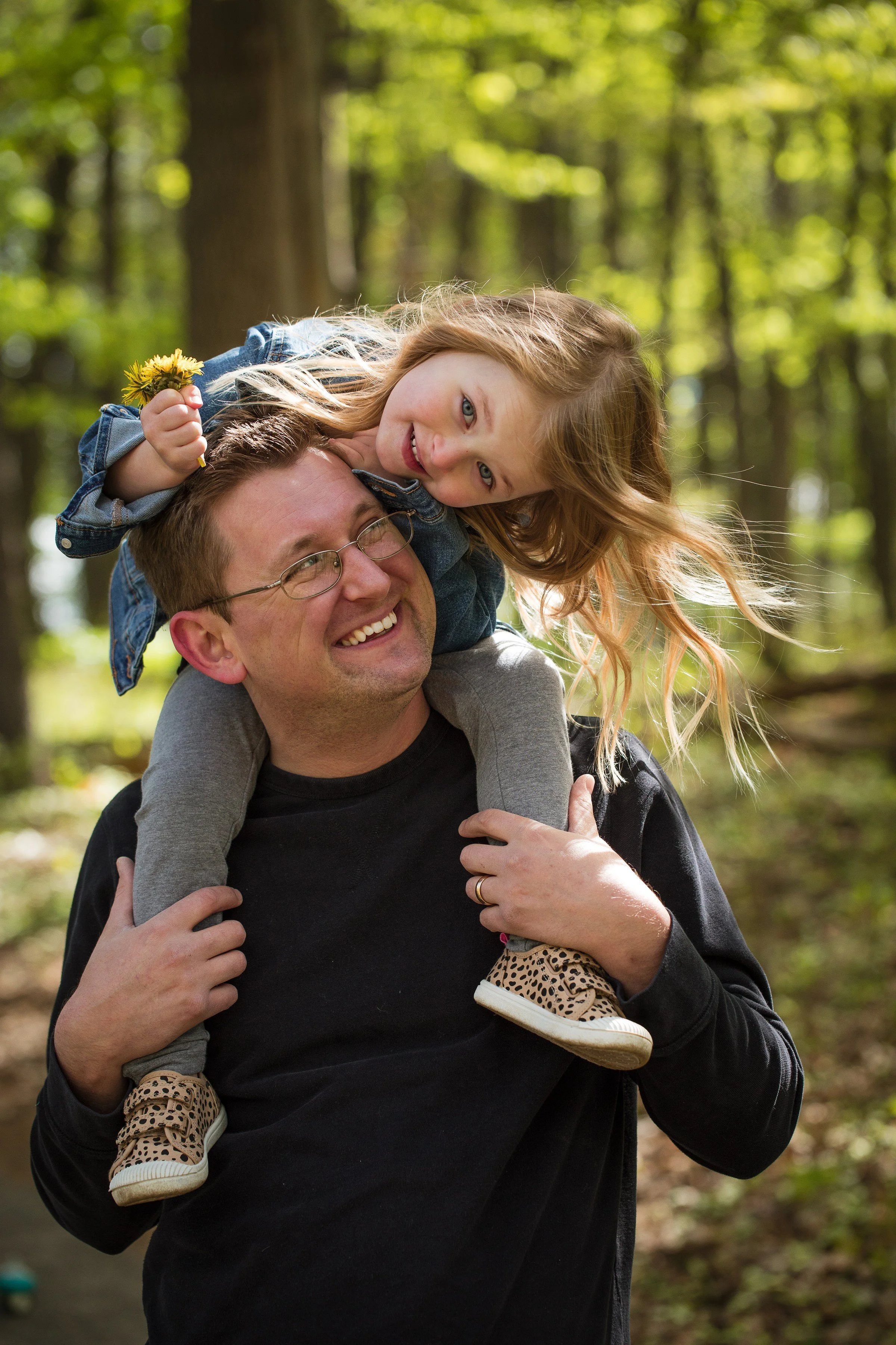 A man with glasses carrying a young girl on his shoulders in a wooded area.