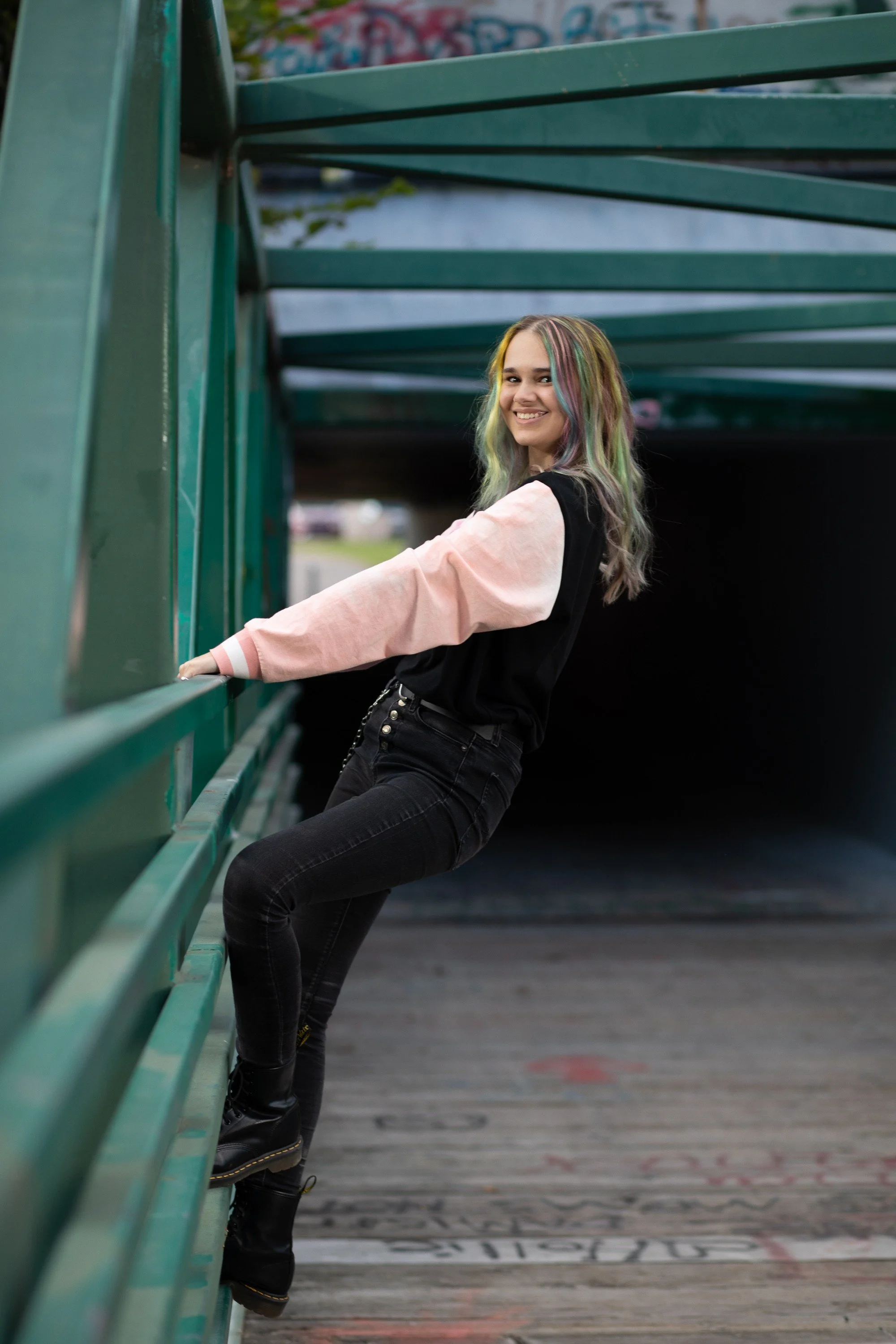 A young woman with colorful, wavy hair smiling and leaning on a green metal railing at an outdoor skate park.