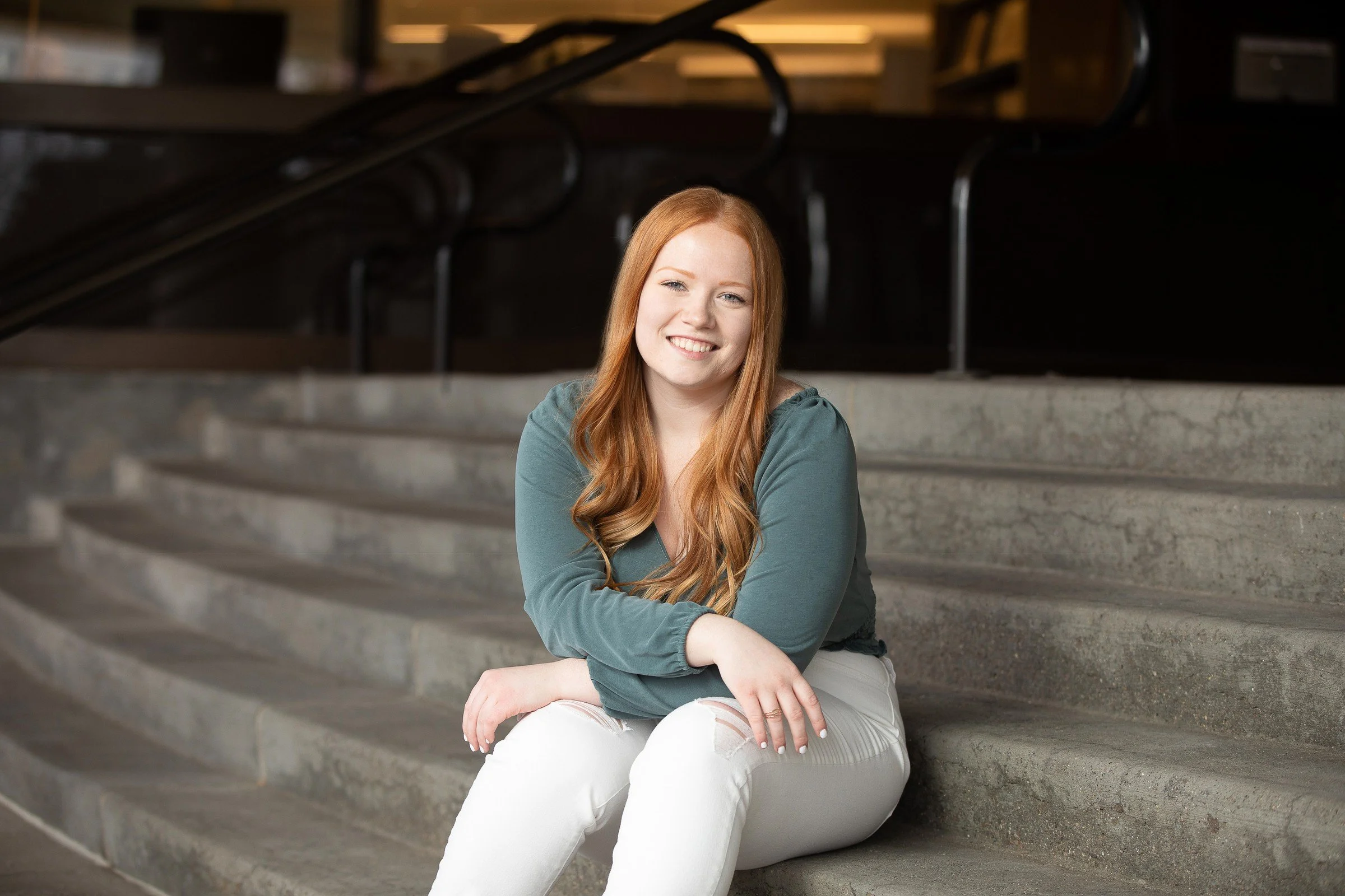 A young woman with red hair, sitting on concrete stairs, smiling at the camera in an indoor setting.