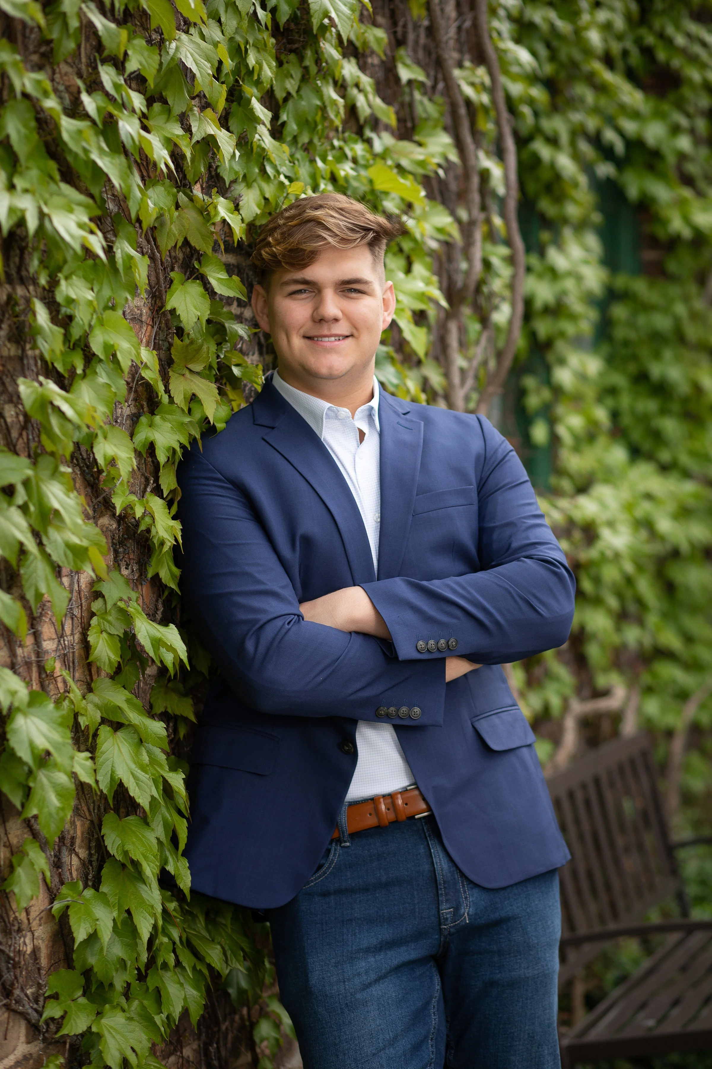 Young man in a blue blazer and jeans, standing with arms crossed and smiling, leaning against a wall covered in green ivy.