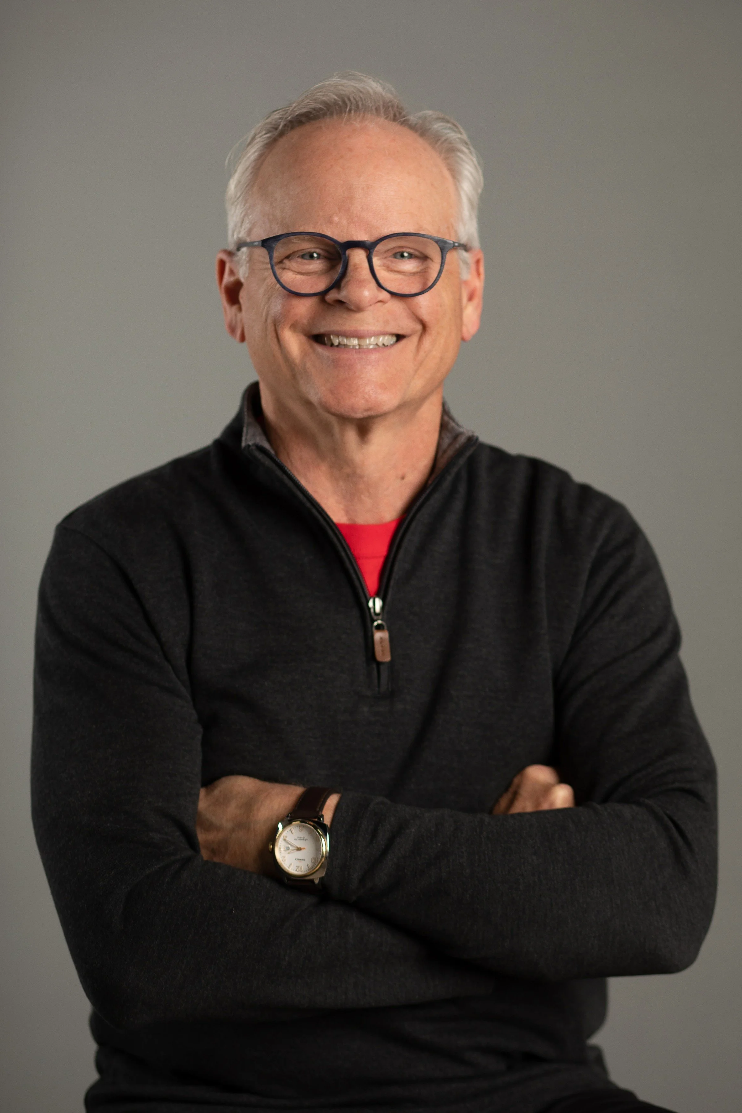 A smiling man with gray hair, glasses, a black zip-up sweater, a red shirt underneath, and a watch on his left wrist, standing against a gray background.