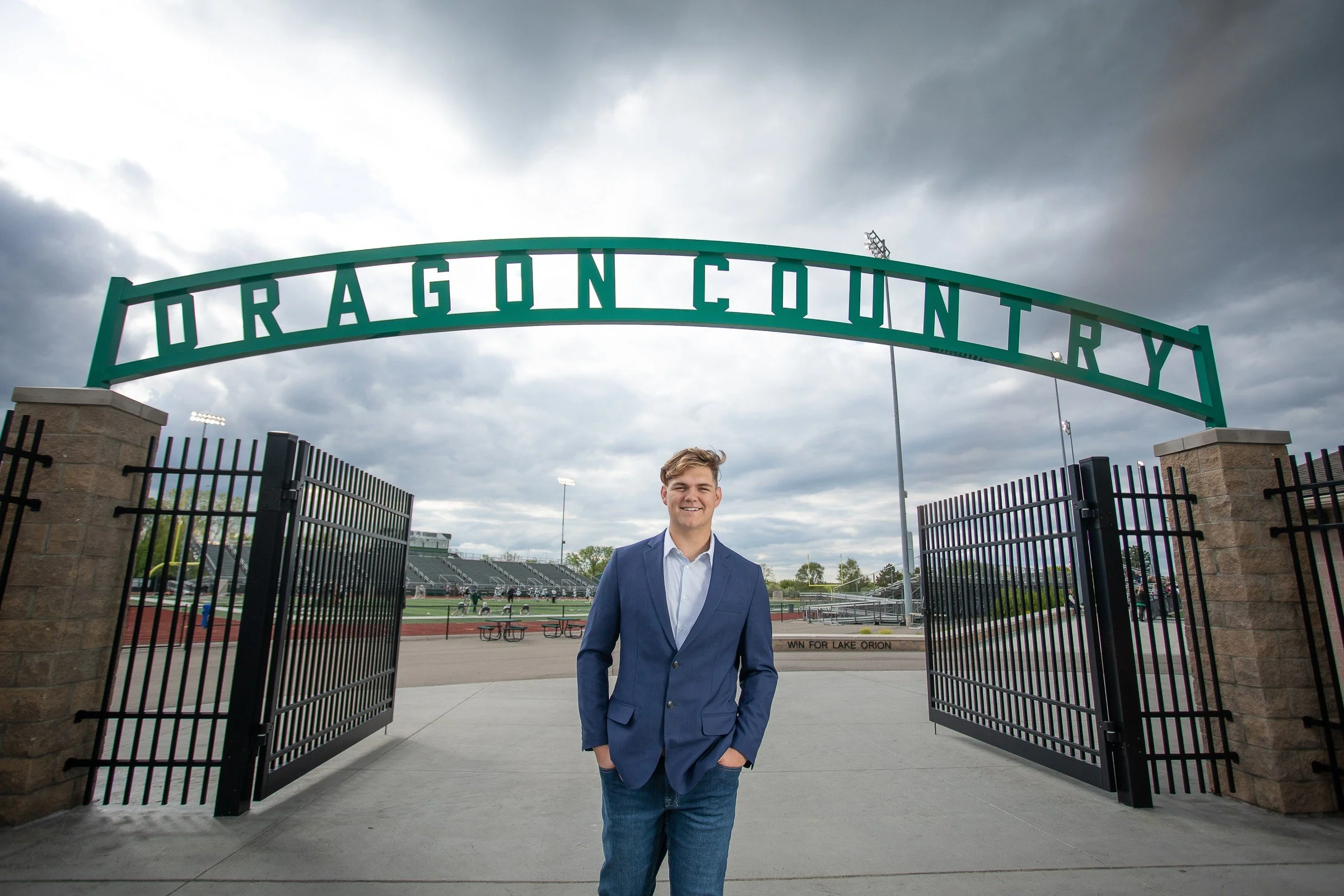 A young man in a blue blazer and jeans standing at the entrance of Dragon Country stadium, with an overcast sky and stadium seating in the background.