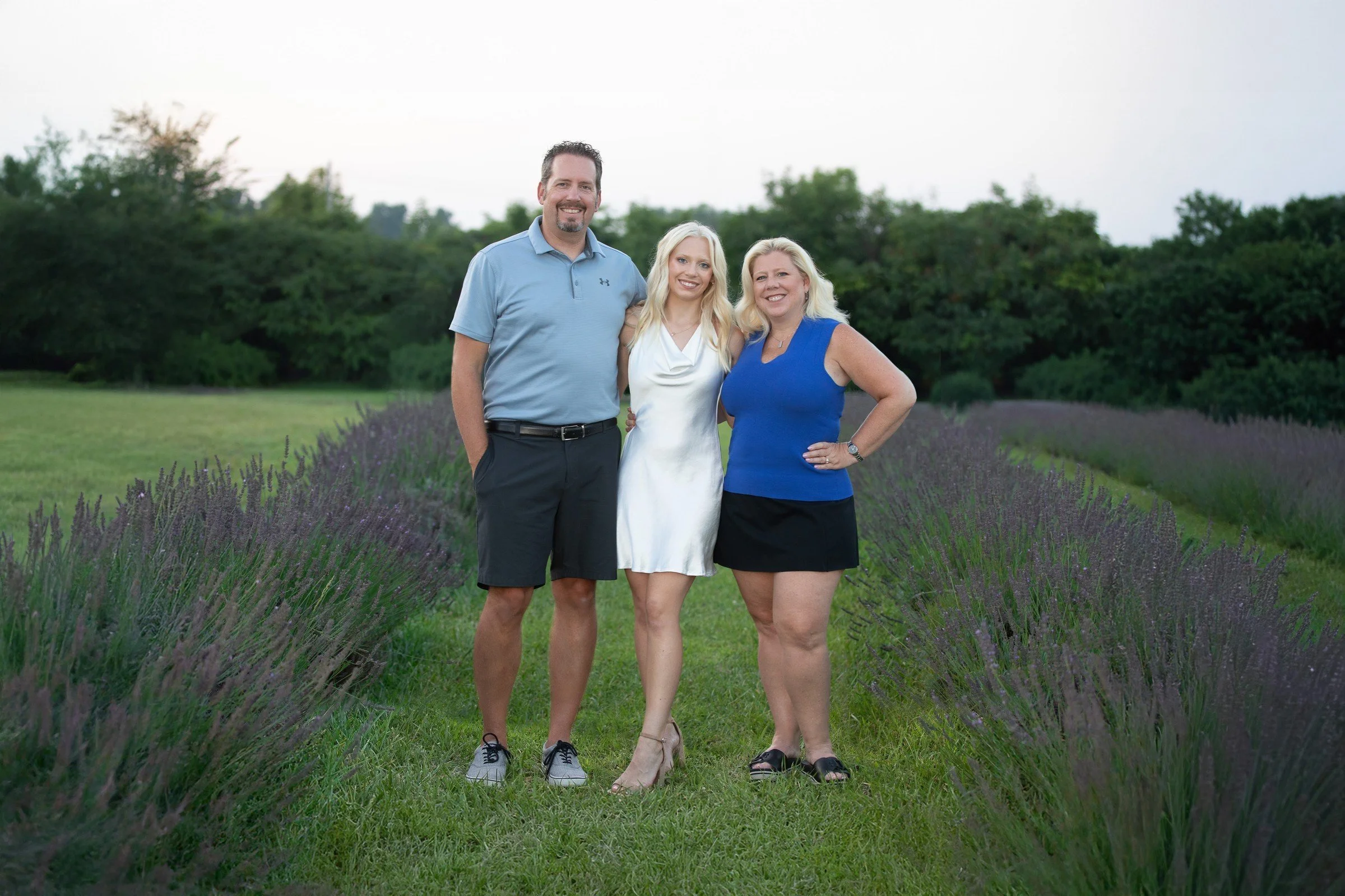 Three people standing together outdoors on a grassy path, surrounded by lavender plants and trees in the background, during daytime.