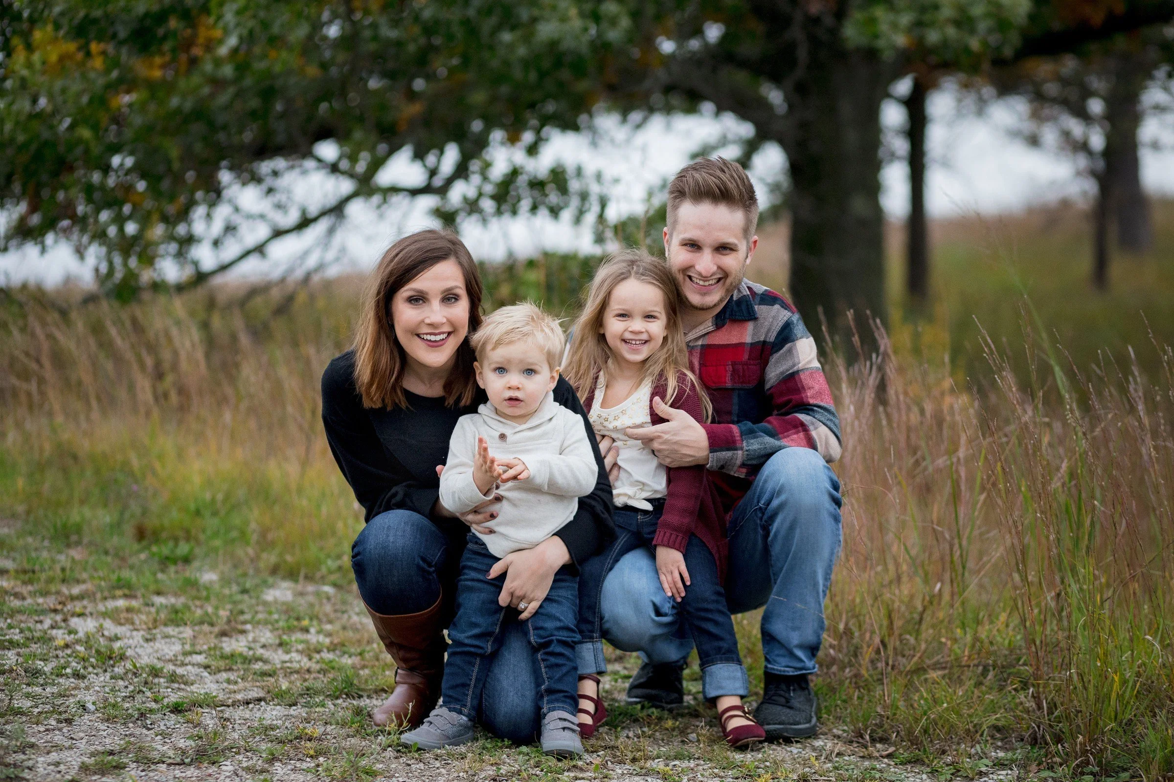 A family of four outdoors on a grassy and gravel path, smiling at the camera. The mother and father are kneeling with their two children, a boy and a girl, in a park with trees and grass in the background.