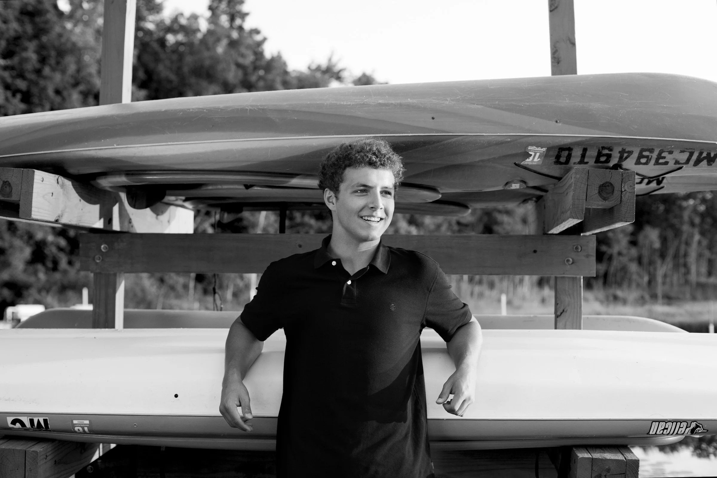 A young man with curly hair smiling, standing next to a kayak and surfboards stored on a wooden rack outdoors.