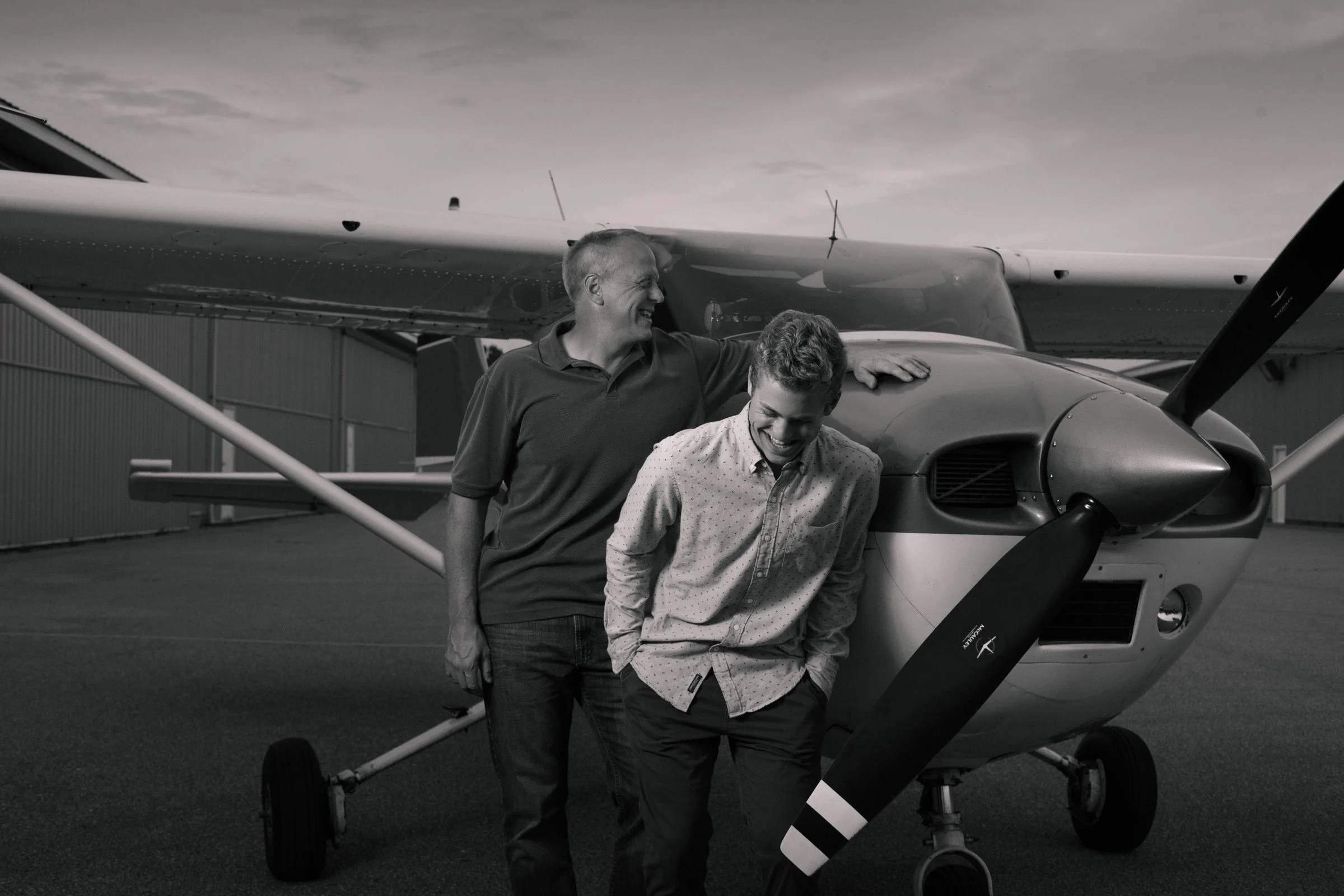 Two men standing in front of a small airplane on an airfield, smiling and sharing a light moment, black and white photo.