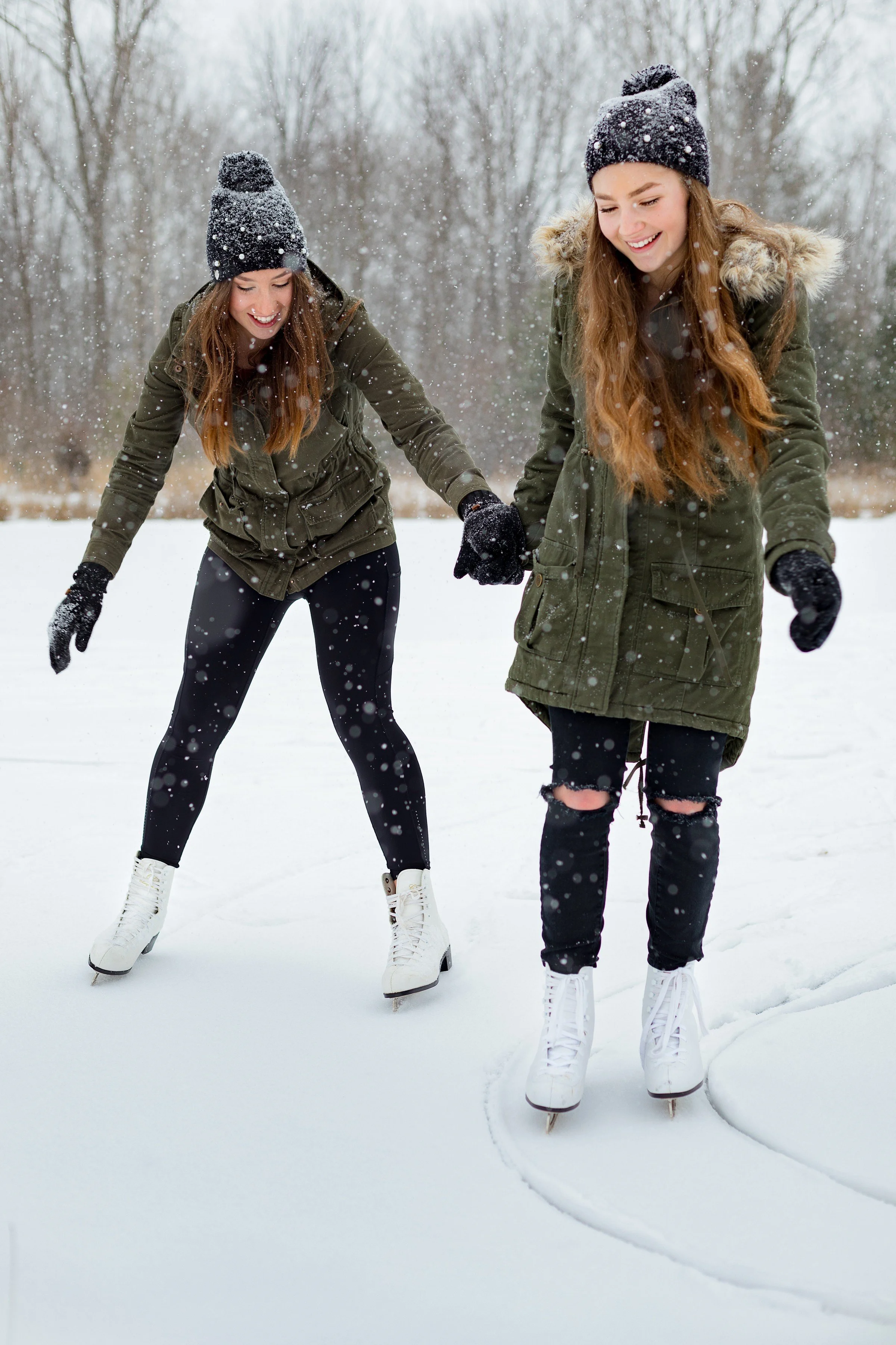 Two young women ice skating outdoors in a snowy winter landscape, wearing warm jackets, beanies, and gloves, enjoying winter activity.