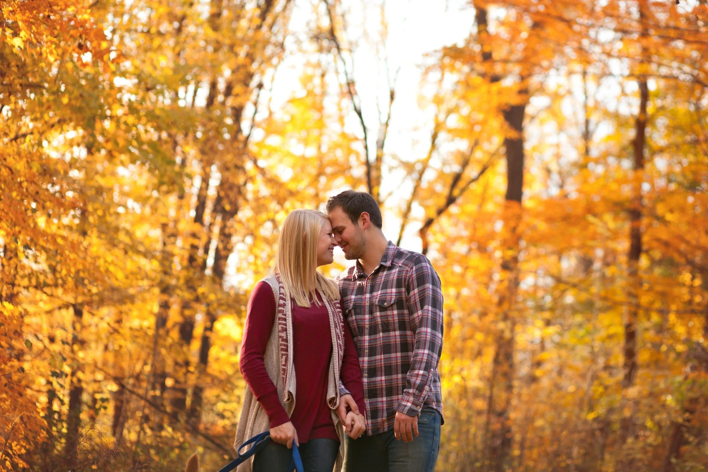 A couple holding hands standing close together in a forest with autumn leaves, smiling and touching foreheads.