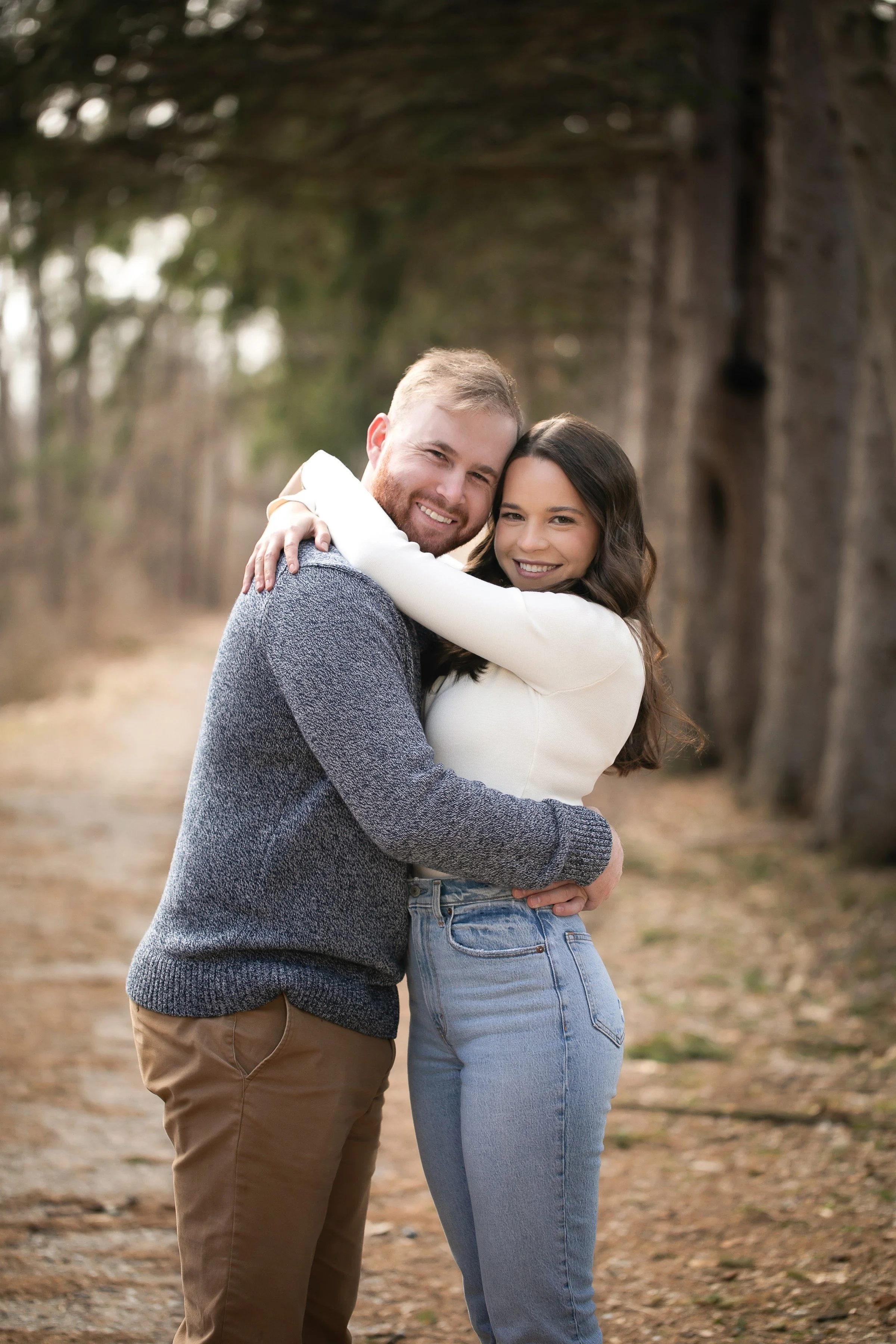 A smiling couple hugging outdoors on a dirt path with trees in the background.