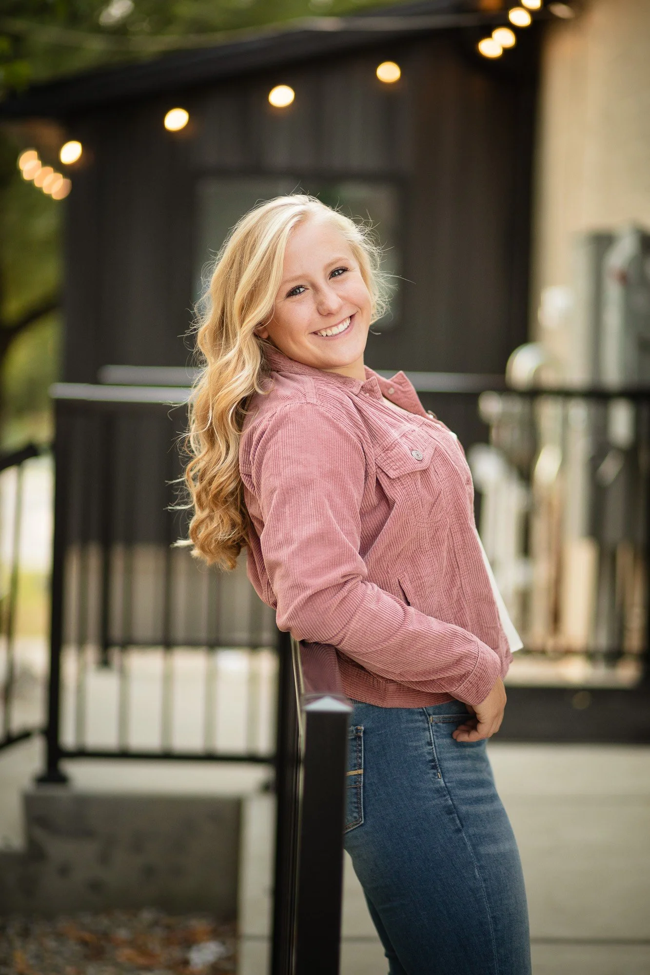 A young woman with long blonde hair smiling and leaning against a black railing outdoors, with string lights overhead and a dark building in the background.