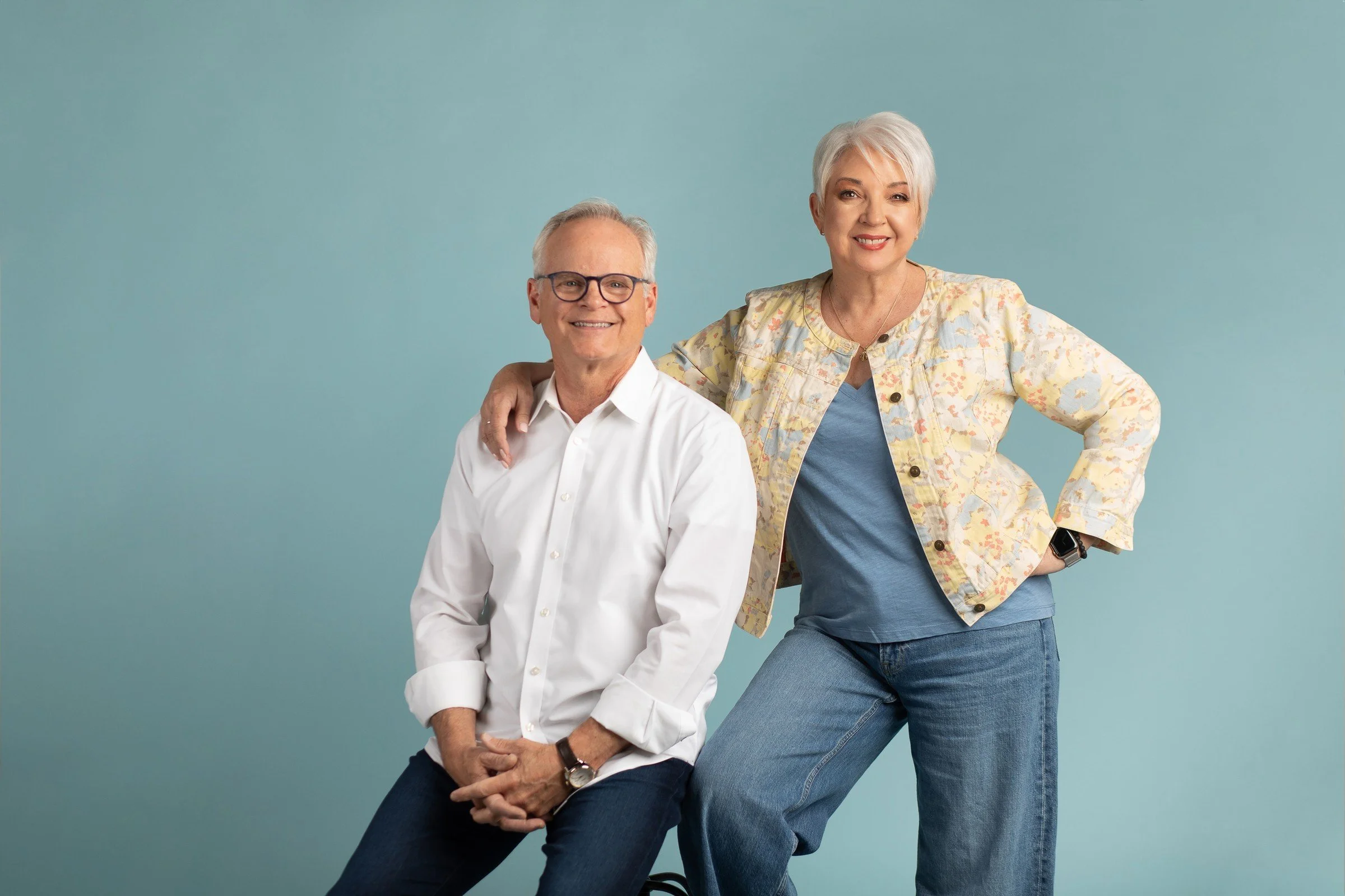 An older man with glasses and short gray hair sits on a stool, smiling, wearing a white button-up shirt and dark jeans. An older woman with short white hair stands next to him, smiling, wearing a floral blazer, a blue shirt, and blue jeans, with one 