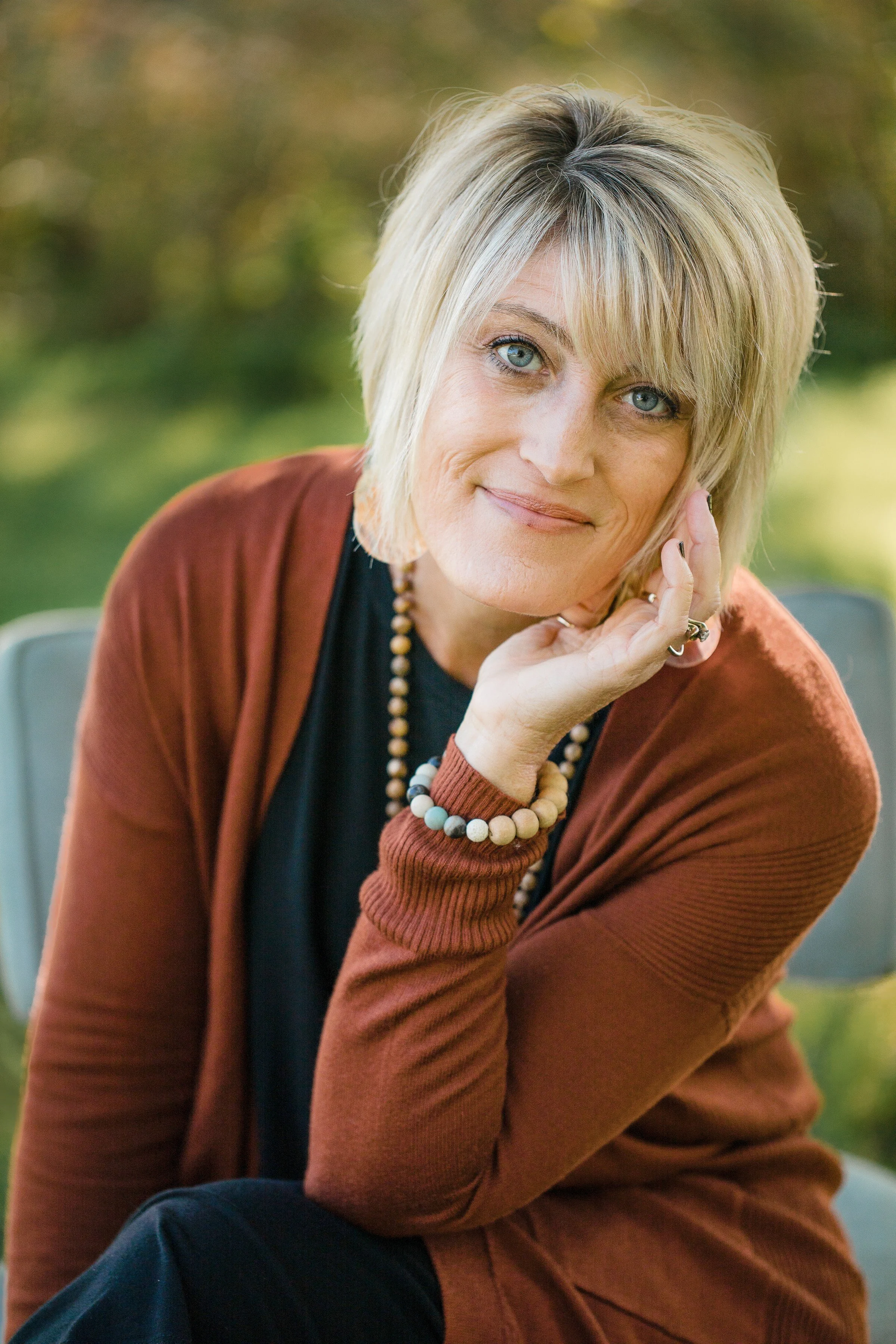 A woman with blonde hair, blue eyes, in a black top and rust-colored cardigan, sitting outdoors with green blurred background, resting her chin on her hand, looking at the camera.