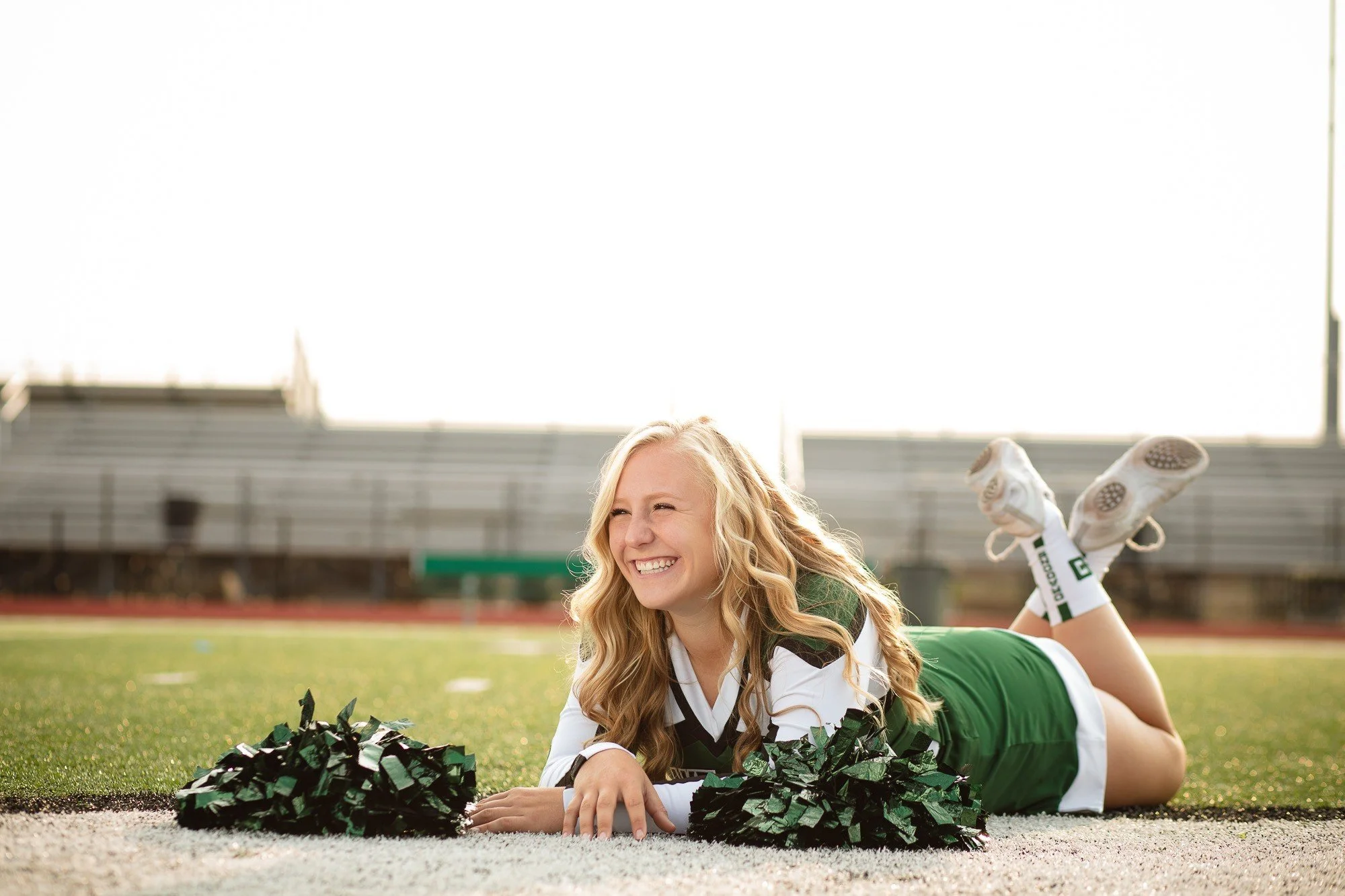 A cheerful young woman in a football uniform lying on her stomach on a football field with pom-poms, smiling and looking to the side.
