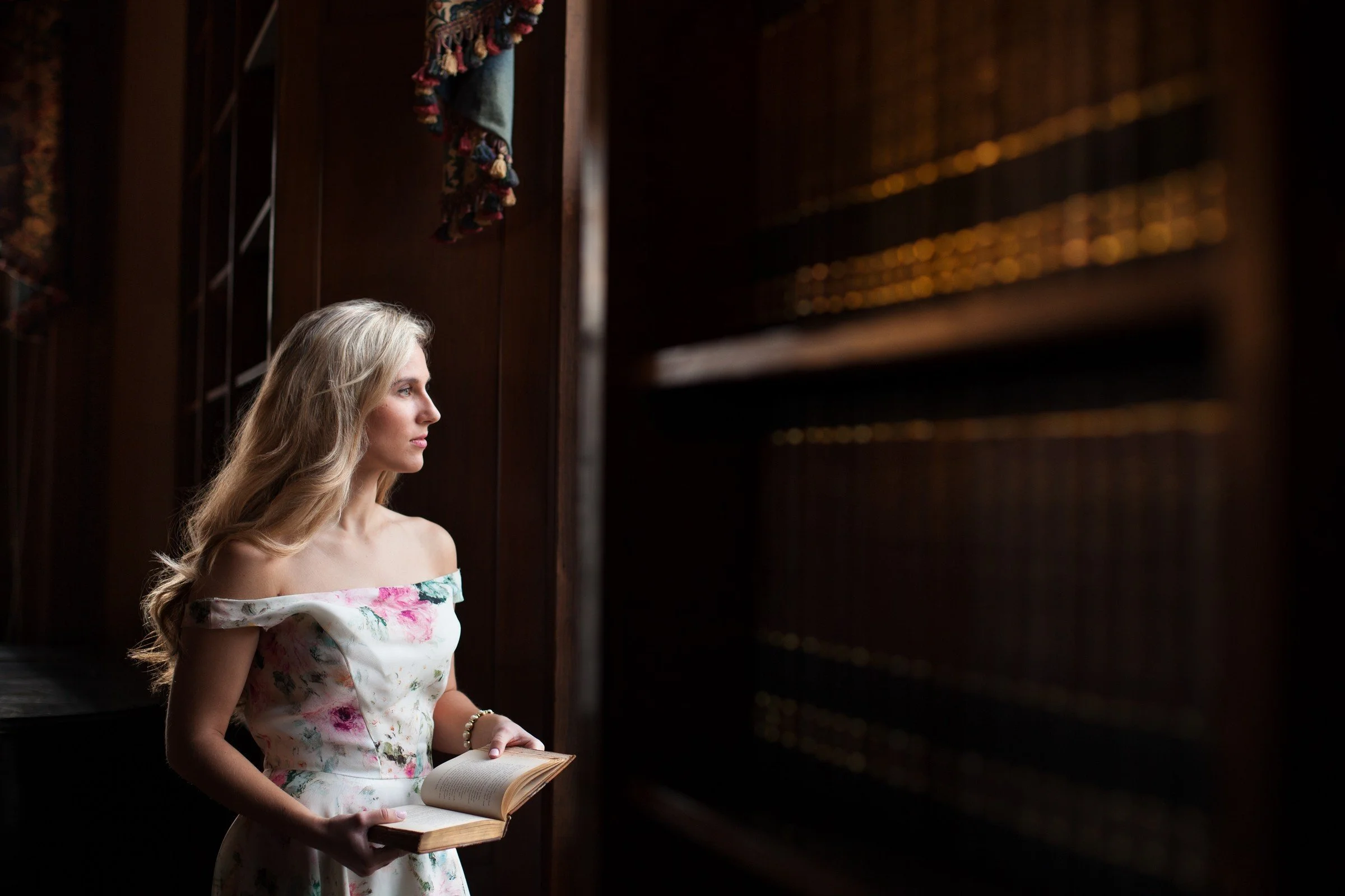 A young woman with long blonde hair standing in a dimly lit library, holding an open book, looking to her left.