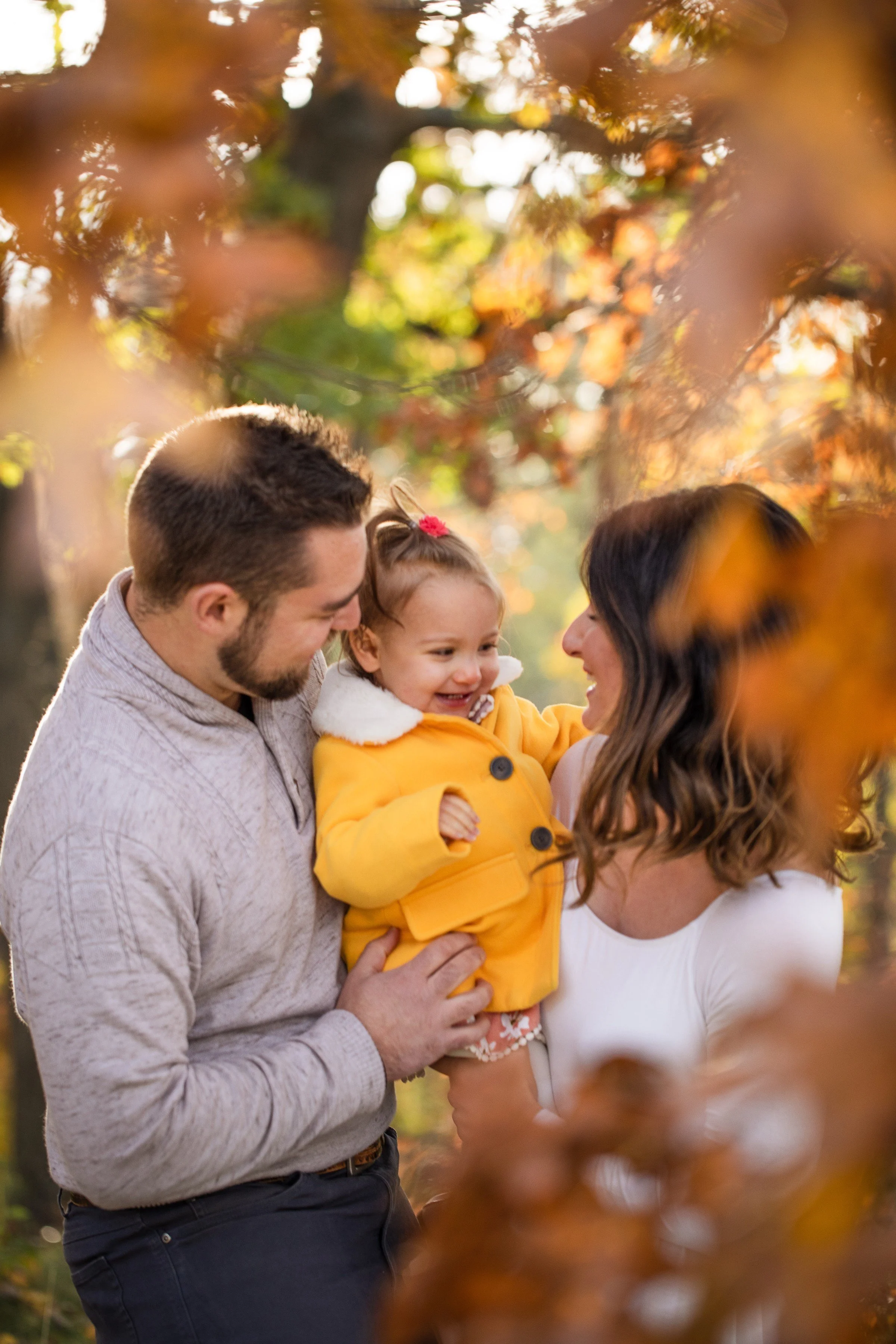 Family of three celebrating fall outdoors, surrounded by autumn leaves. Father and mother hold their smiling young daughter, who wears a yellow coat.