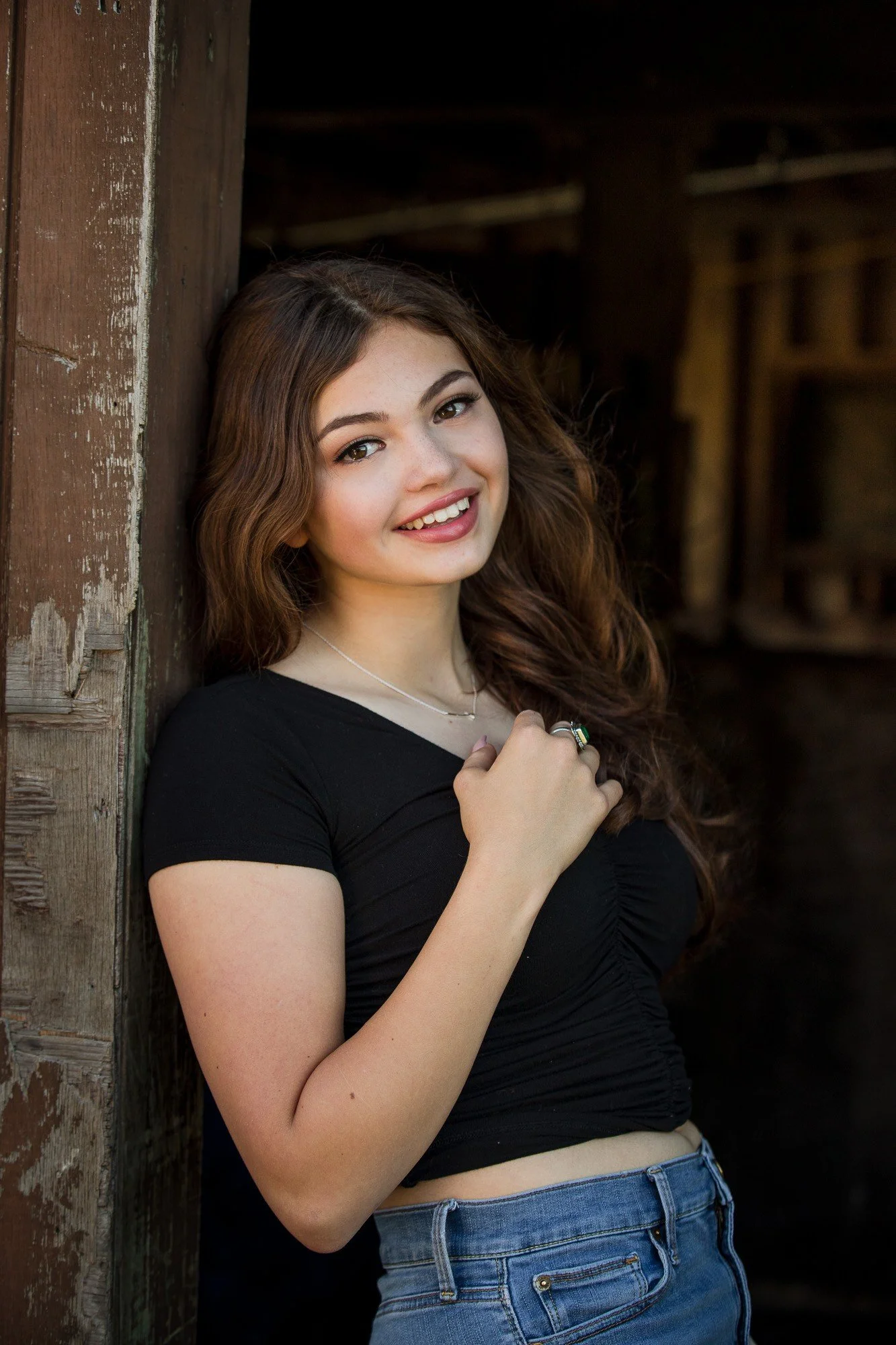 A young woman with long, wavy brown hair and fair skin, smiling and looking at the camera, leaning against a wooden post in a rustic setting, wearing a black top and blue jeans.