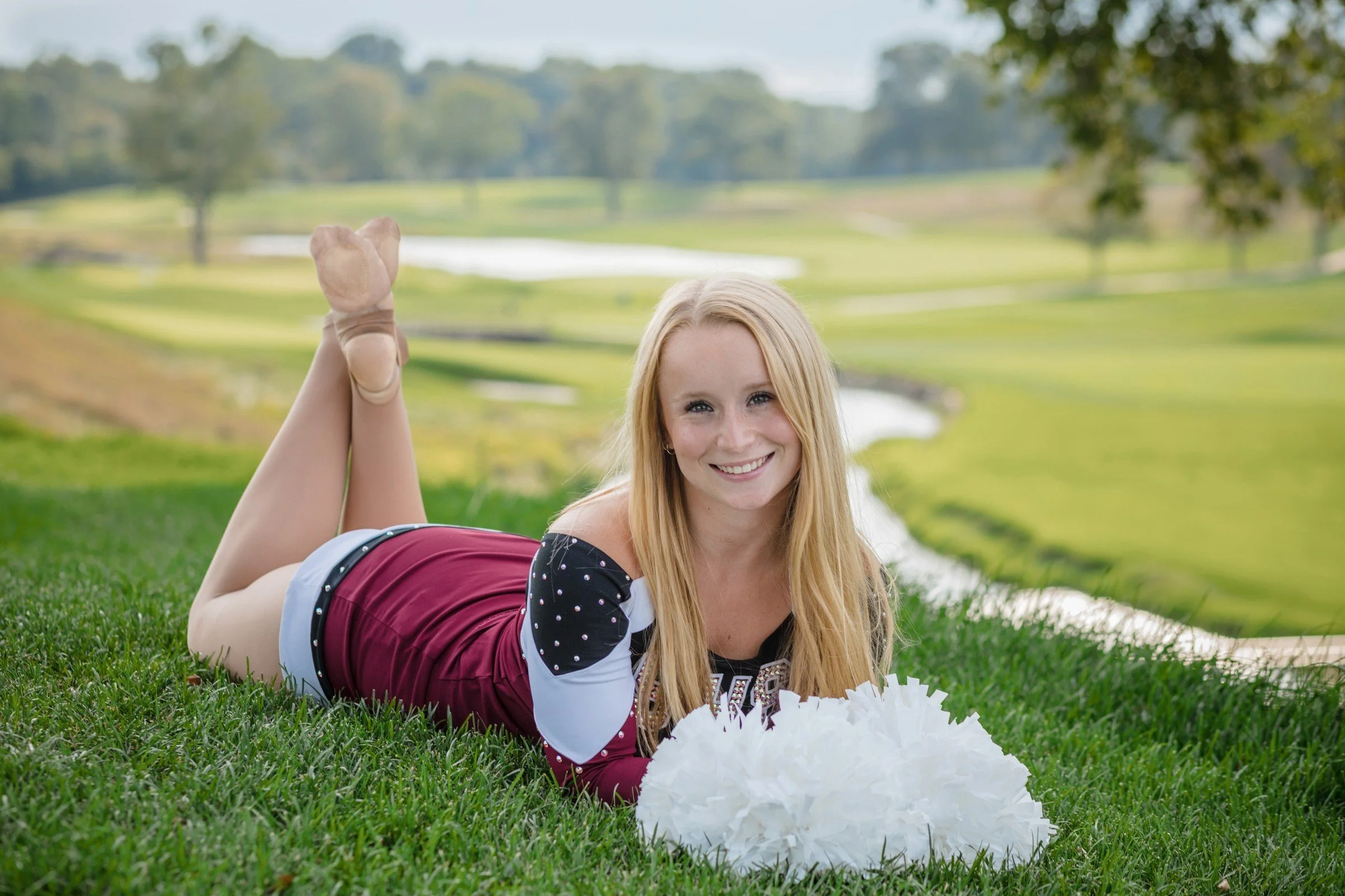 A young woman lying on her stomach on a grassy field, holding white pom-poms, smiling, with a background of a golf course and trees.