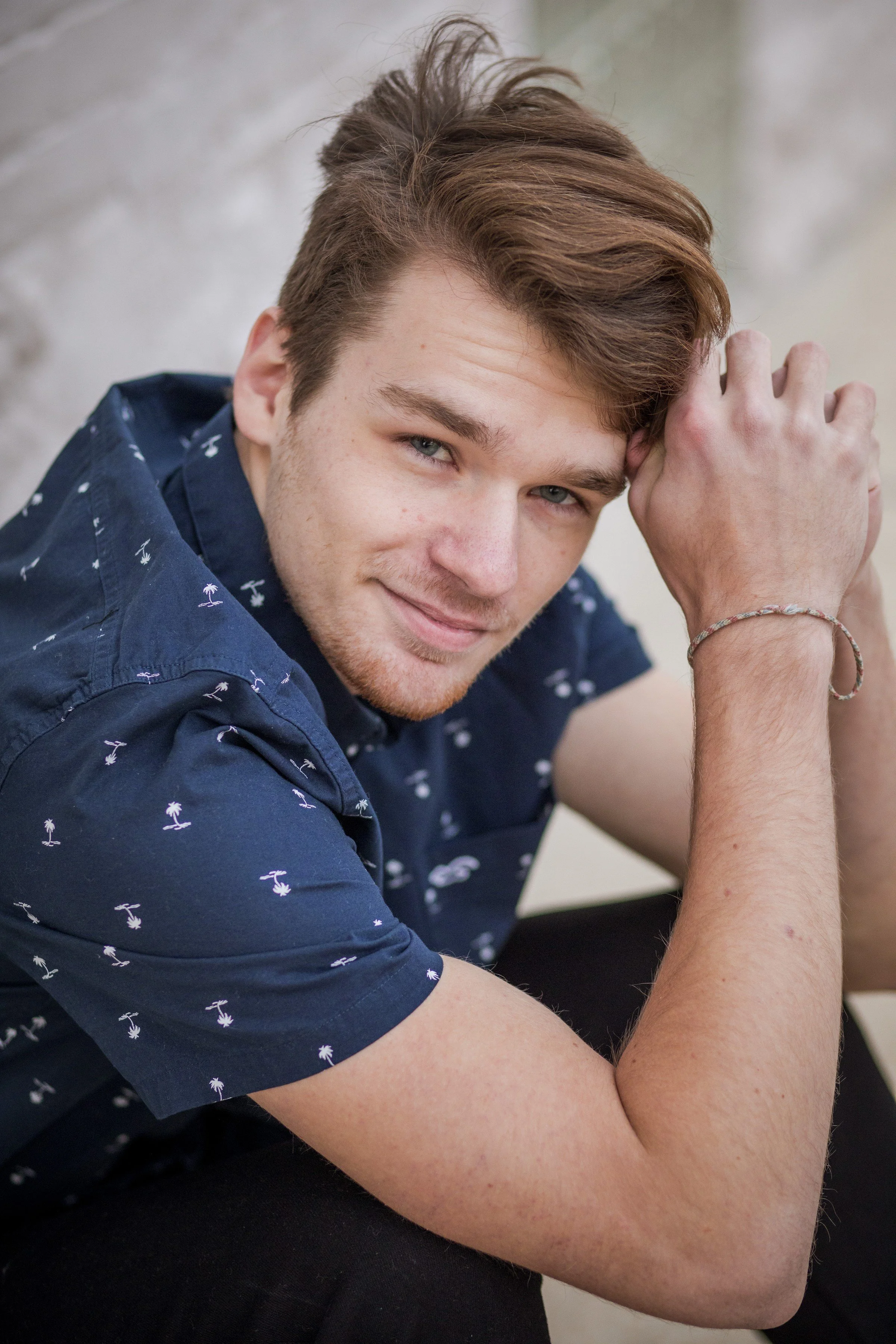 A young man with light skin, short brown hair, and blue eyes, wearing a navy blue shirt with small palm tree patterns, smiling and leaning on his arm against a neutral background.
