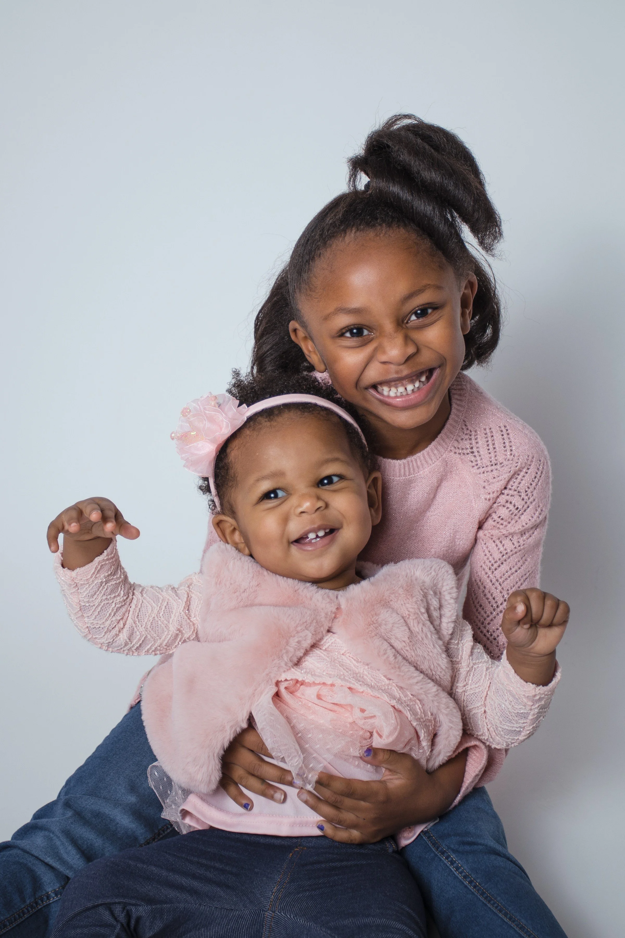 A happy young girl and a toddler girl smiling and posing together, sitting against a plain wall, wearing pink outfits.
