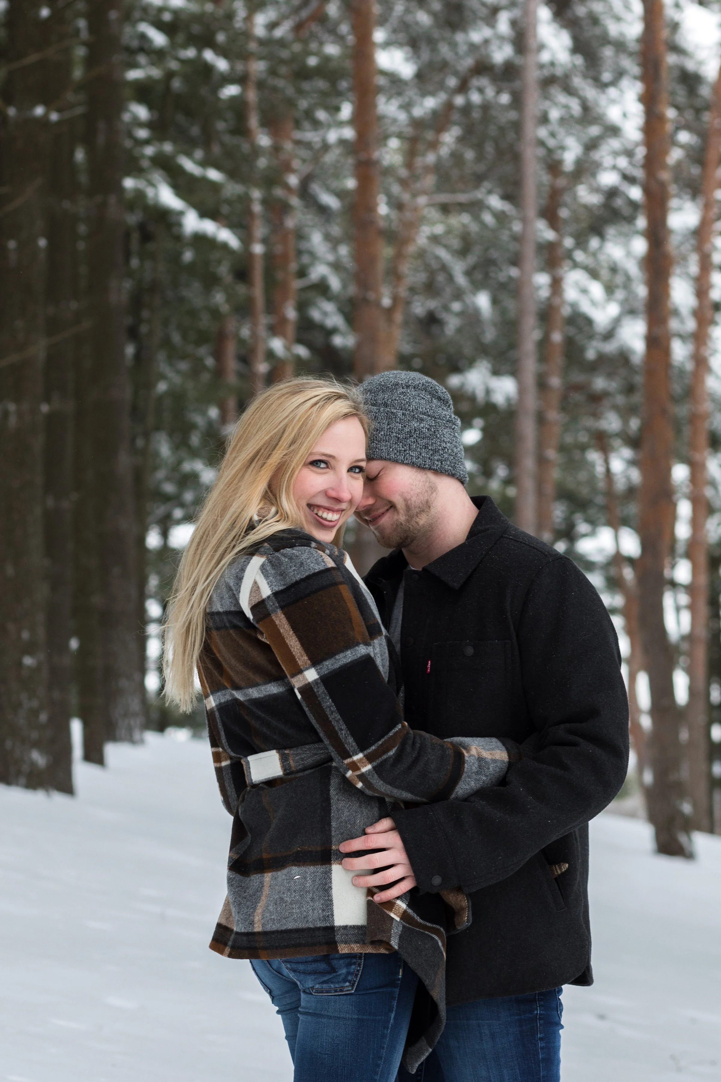A couple standing close together outdoors in a snowy forest, smiling and sharing an intimate moment.