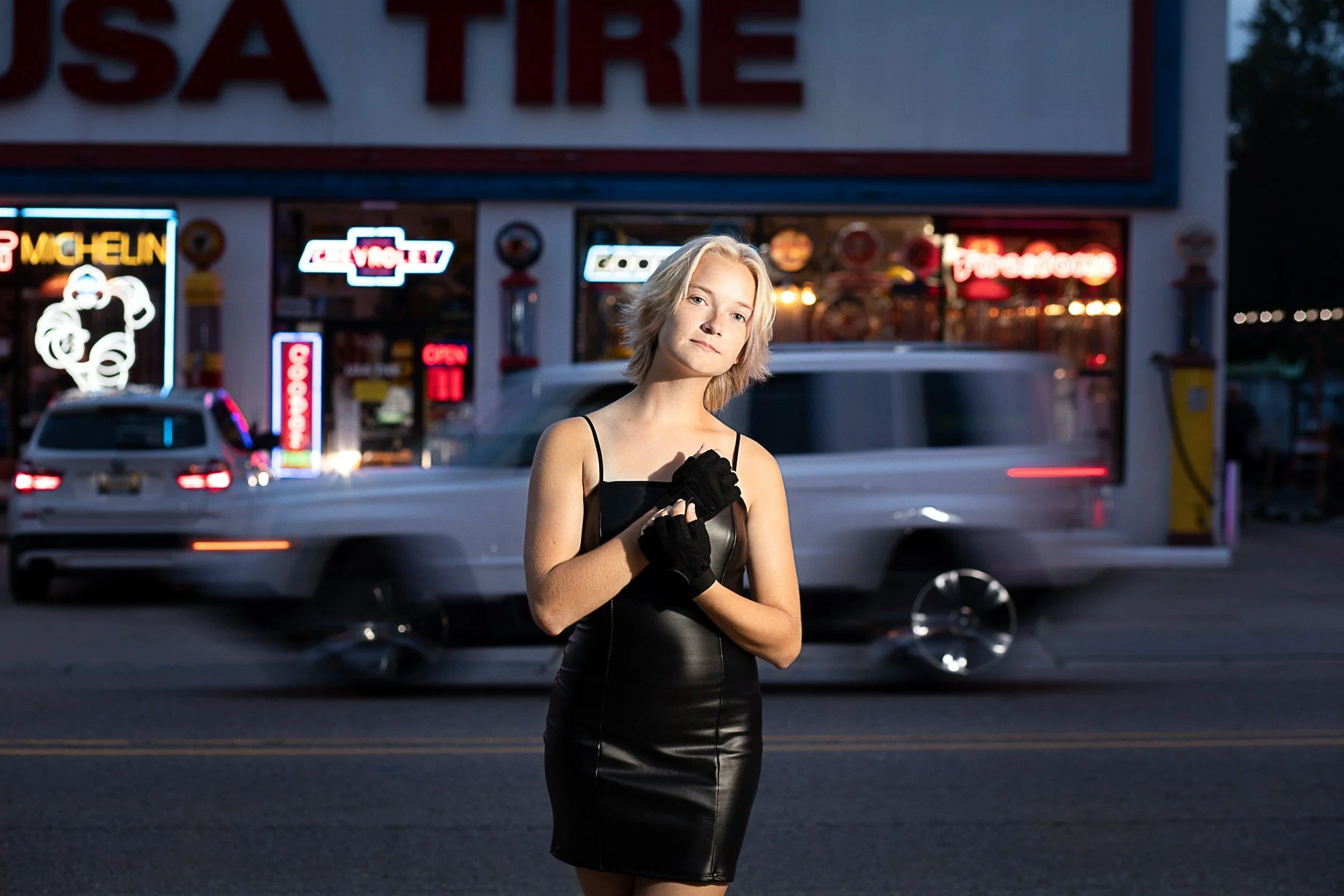 A young woman with short blonde hair stands on a city street at dusk, wearing a black leather dress and black gloves, with neon signs and a moving vehicle in the background.