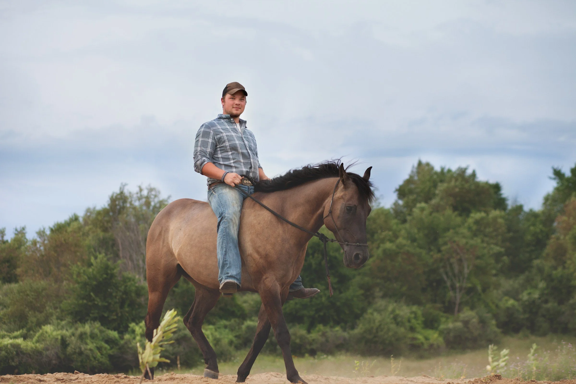 A man riding a brown horse outdoors, with greenery and trees in the background, under a cloudy sky.