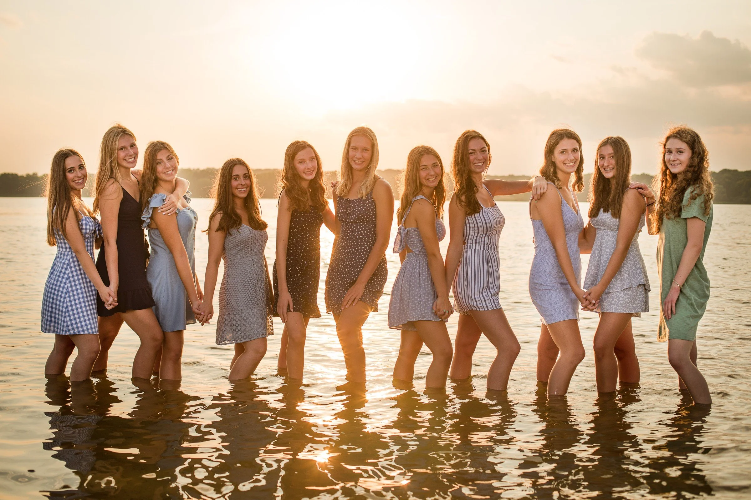 A group of fifteen young women in dresses holding hands in shallow water at sunset, smiling at the camera.