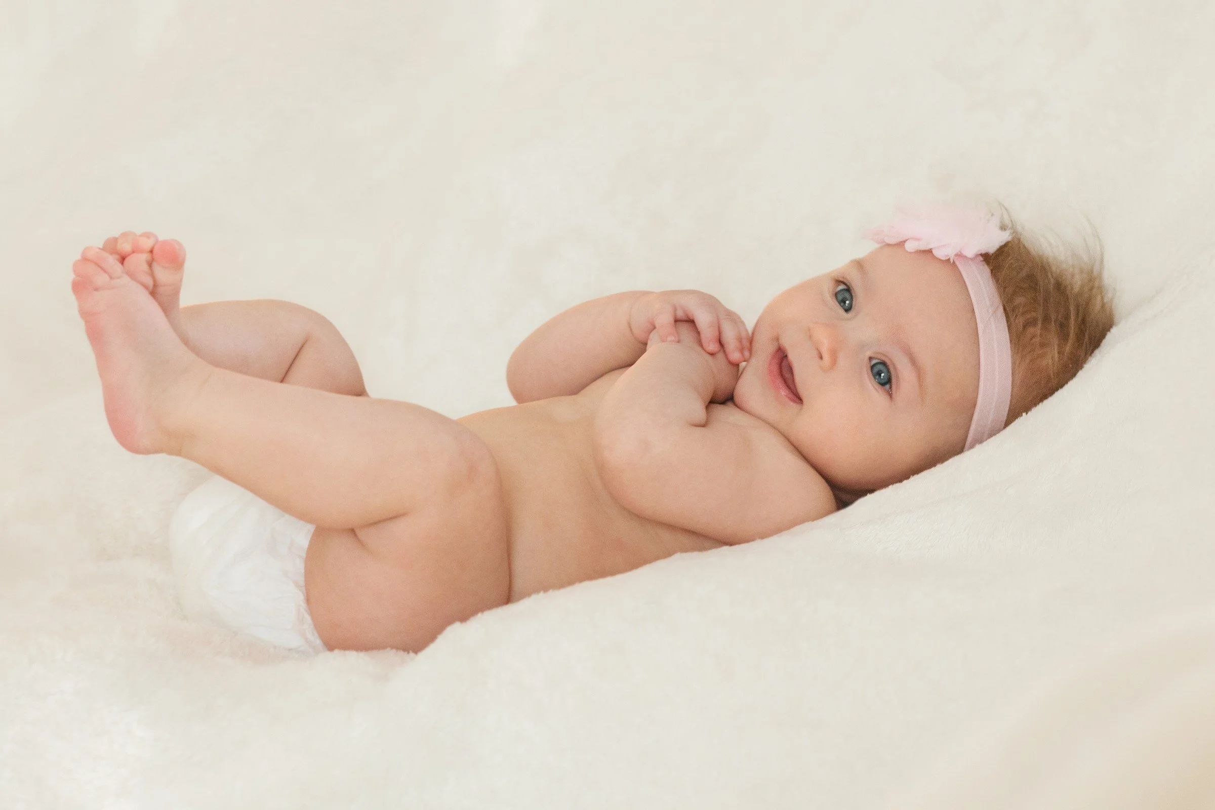 A smiling baby girl with blue eyes wearing a pink headband with a bow, lying on her side on a soft white blanket, with hands near her mouth and legs slightly bent.