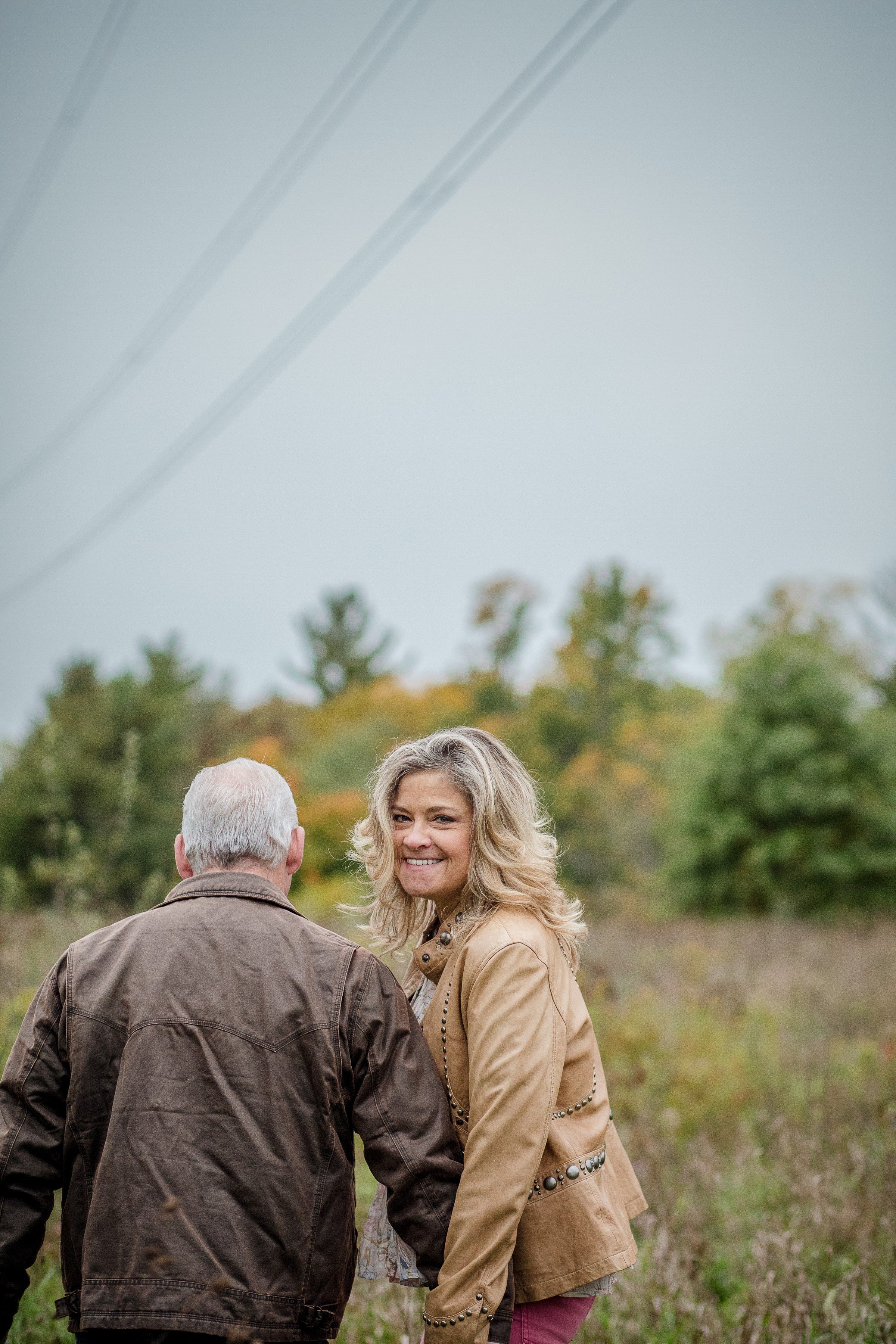 A woman with curly blonde hair smiling at the camera, wearing a tan jacket, in an outdoor setting with trees and overcast sky in the background, standing next to an older man with gray hair, wearing a brown jacket, with their backs partially turned.