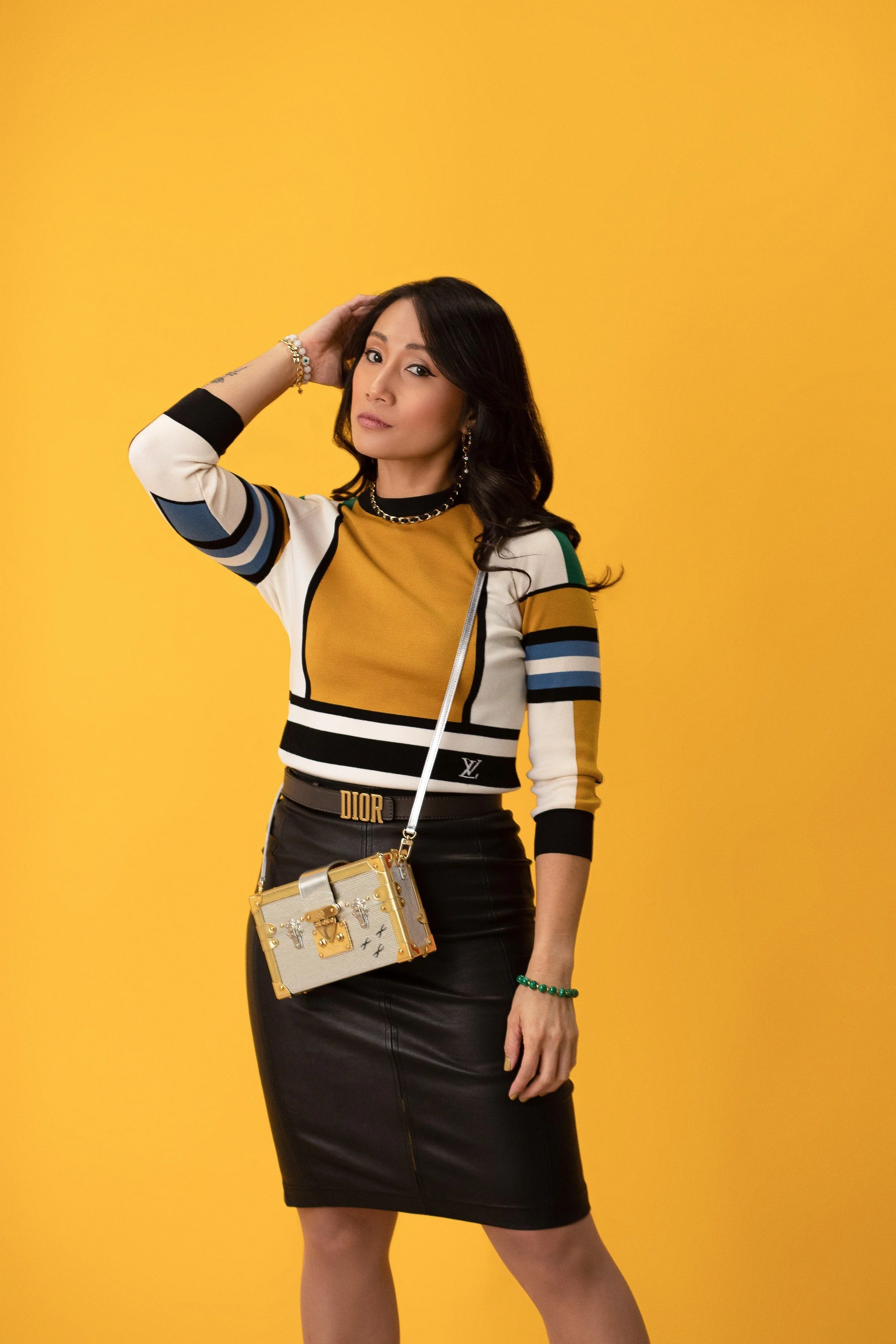 A woman wearing a colorful striped sweater and a black leather skirt, carrying a designer bag, standing against a yellow background.