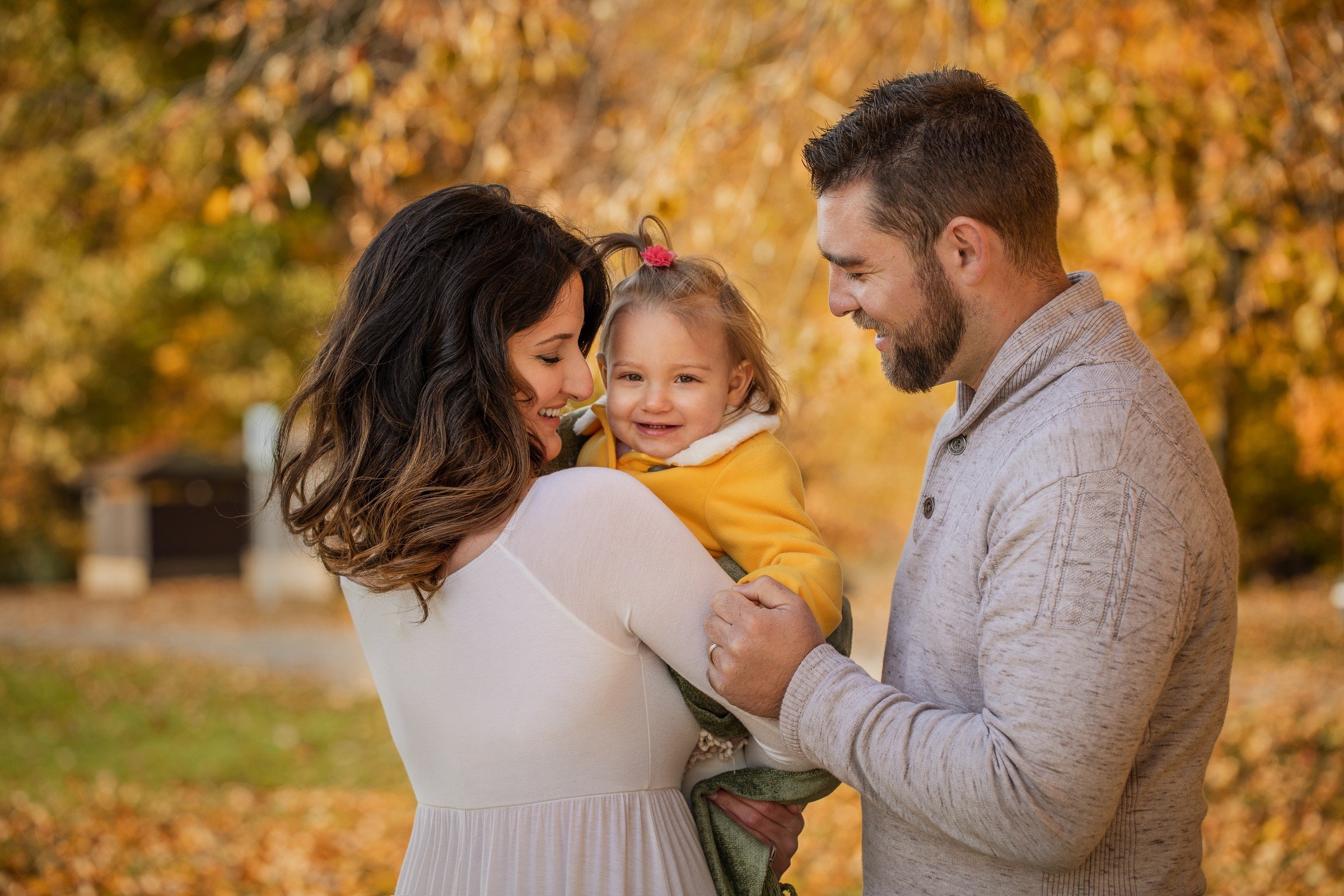 A smiling young family of three, a mother, father, and toddler girl, standing outdoors amid fall foliage, with the mother holding the girl and the father touching her arm.