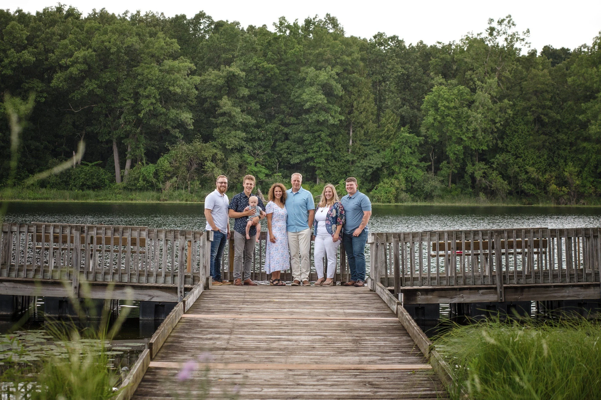 A group of seven people standing on a wooden dock by a lake with lush green trees in the background.