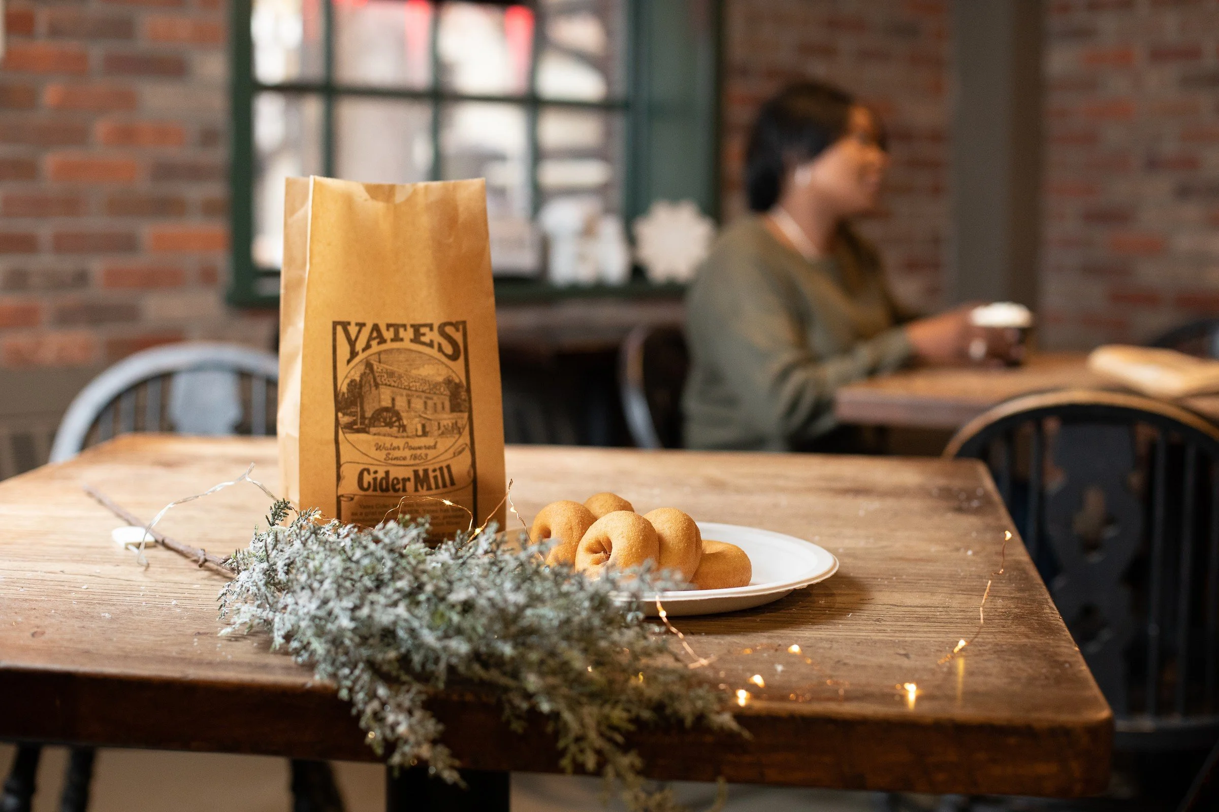 A paper bag labeled 'Yates Cider Mill' on a wooden table, with a plate of small donuts, a sprig of snow-dusted greenery, and string lights, in a cozy cafe setting with a blurred woman sitting by the window in the background.