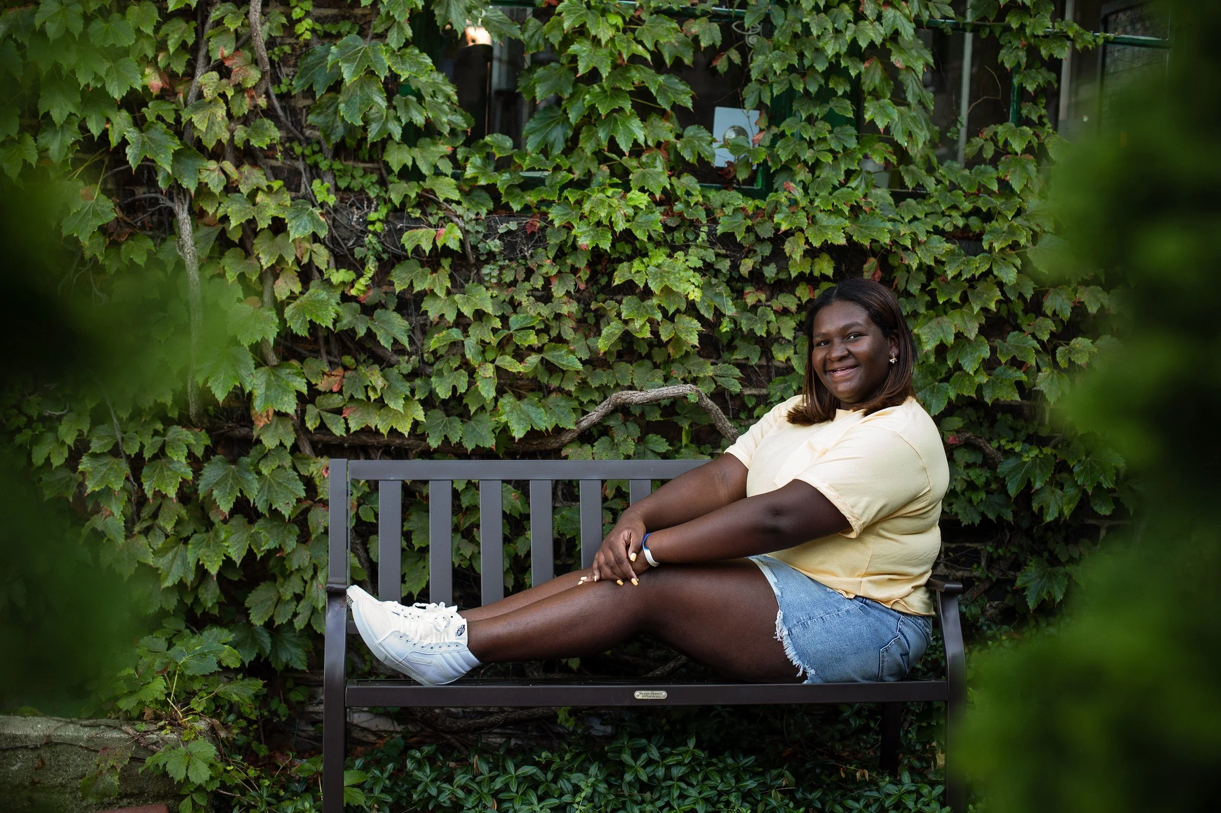 A woman sitting on a bench outdoors, smiling, with green ivy on a wall behind her, wearing a yellow shirt, denim shorts, and white sneakers.