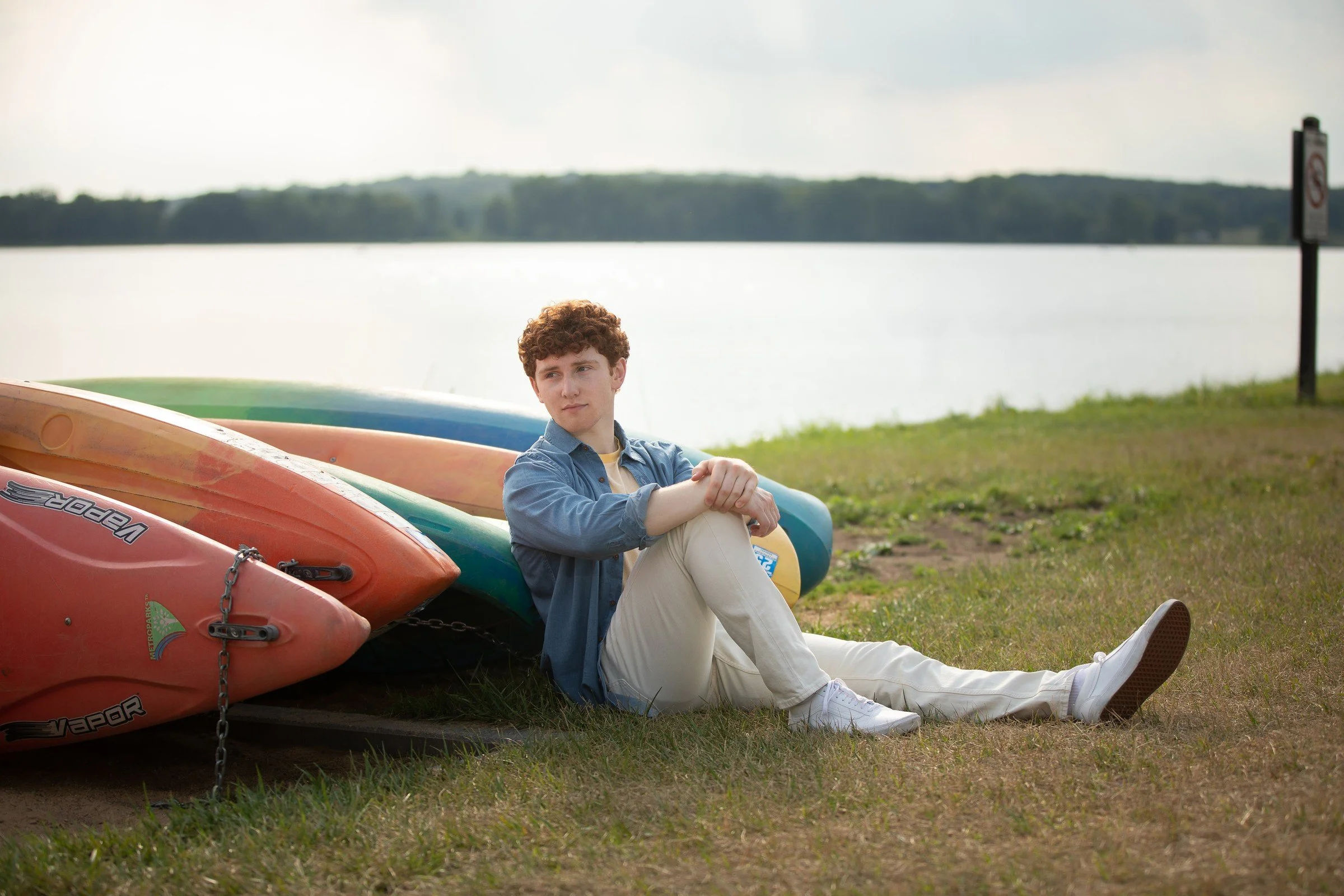 A young man with curly hair sitting on grass near a lake, with multiple kayaks stacked beside him.