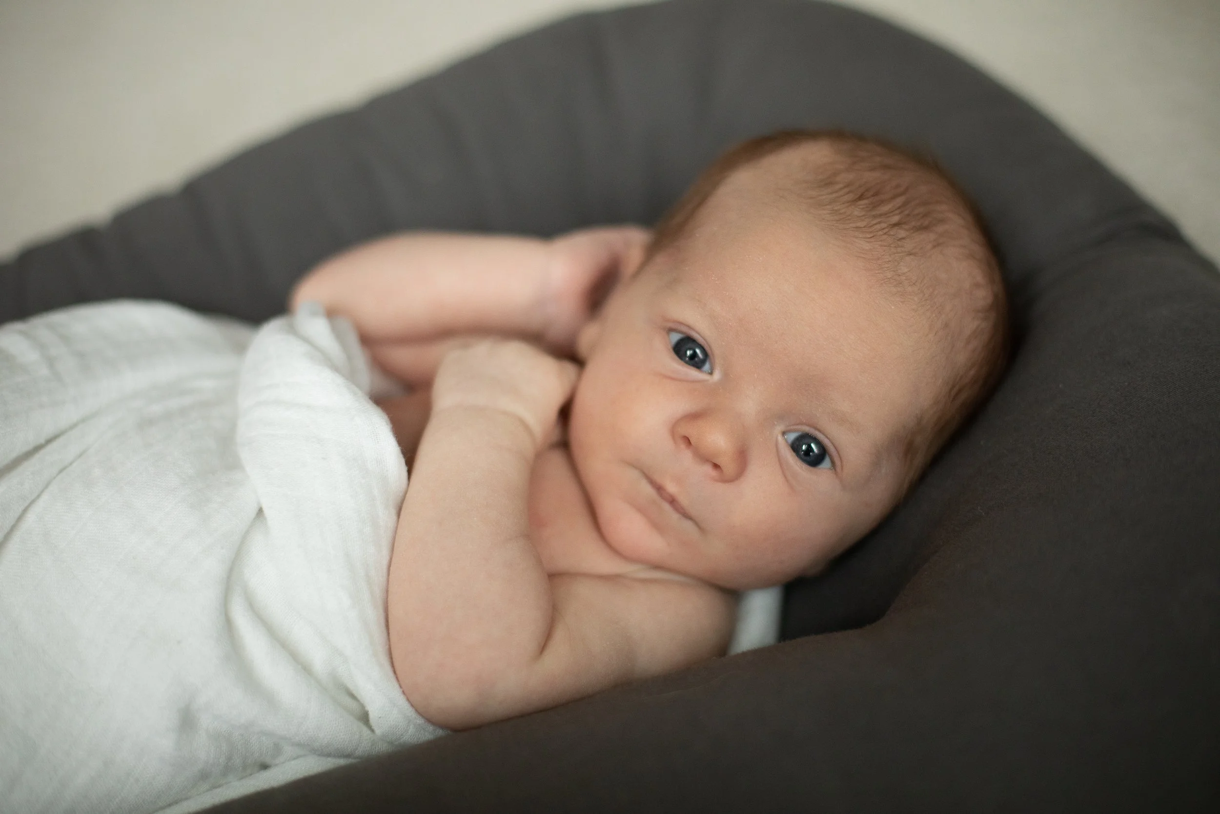 A baby lying on a dark cushion, looking directly at the camera with blue eyes, wearing a light-colored sleeveless top, with one hand near the face and light brown hair.