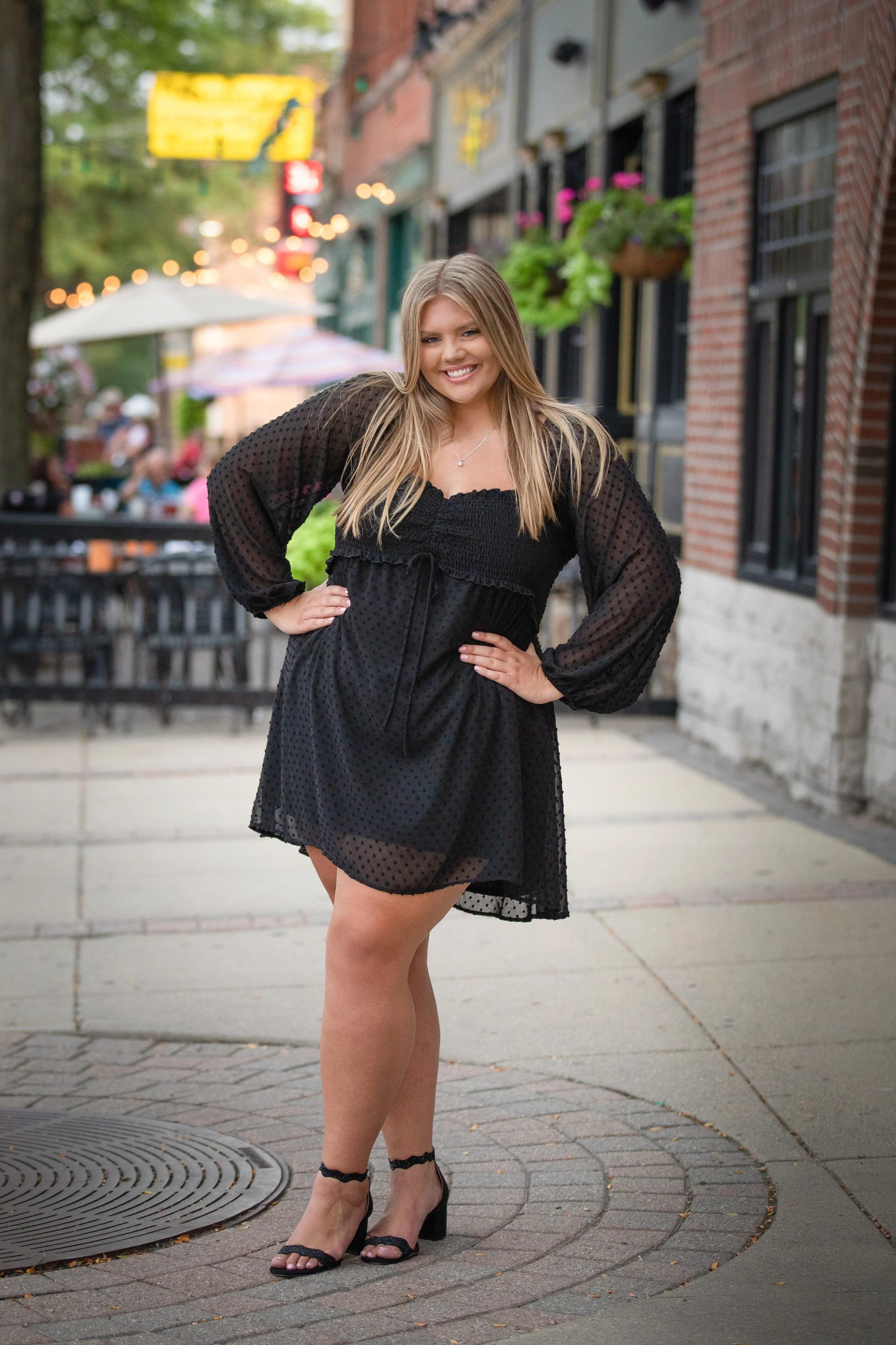 Young woman in a black dress with sheer sleeves posing on a city sidewalk with outdoor seating and buildings in the background.