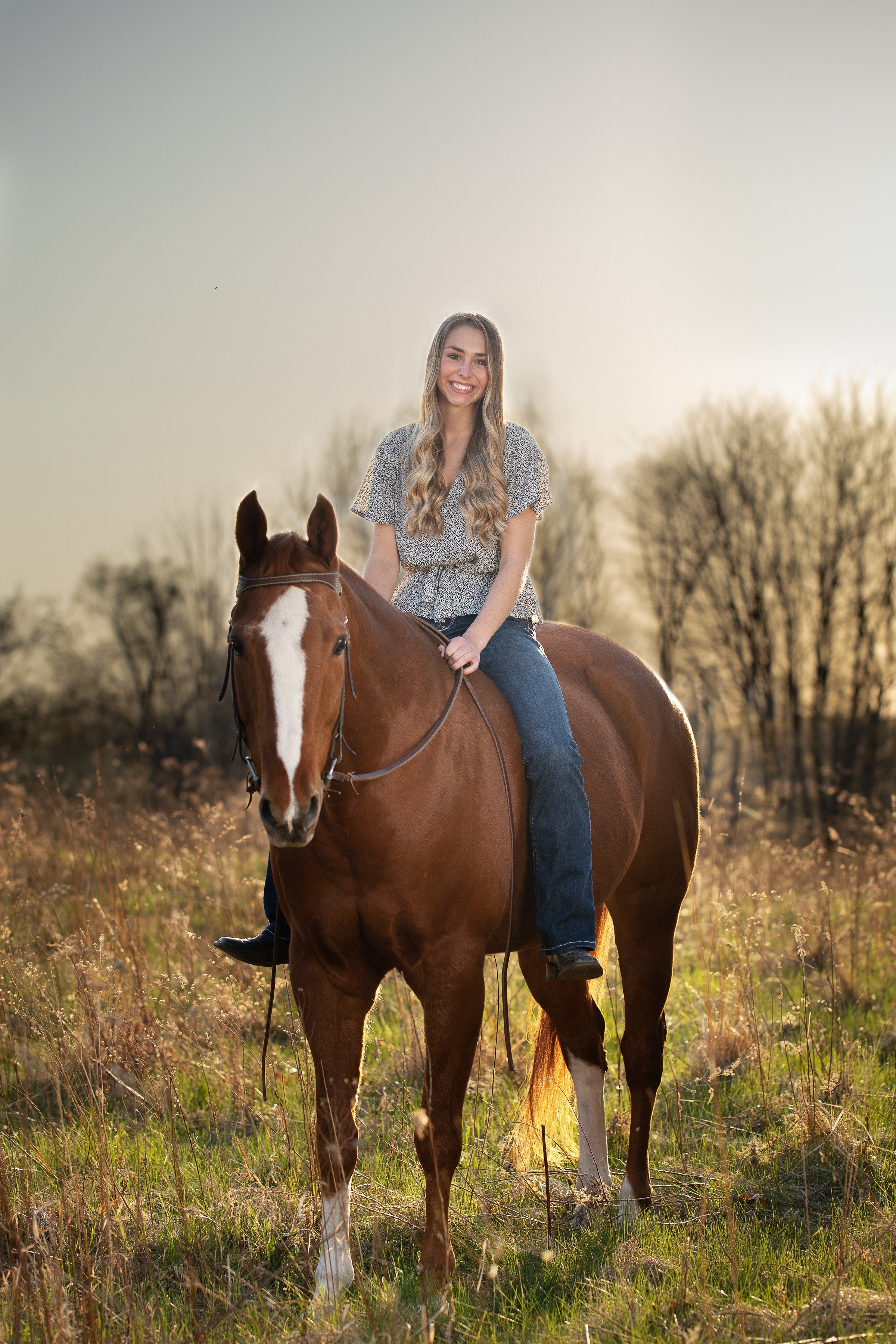 A woman with long wavy hair smiling while riding a brown horse in a grassy field during sunset.