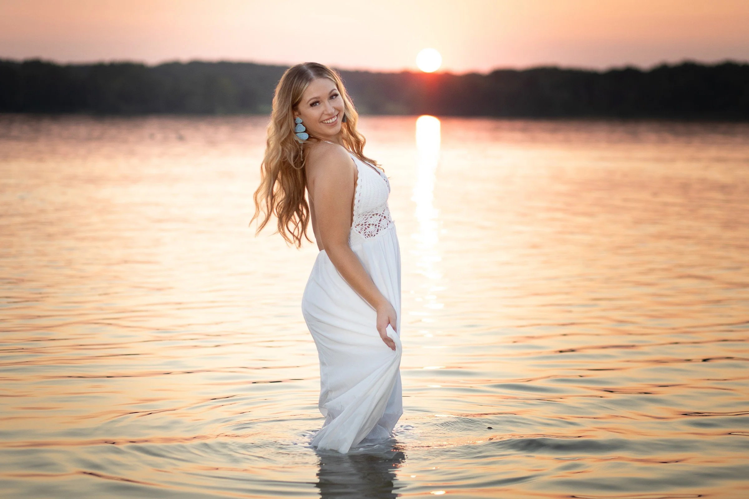 A woman in a white dress standing in a calm body of water at sunset, smiling at the camera with long wavy hair and blue earrings.