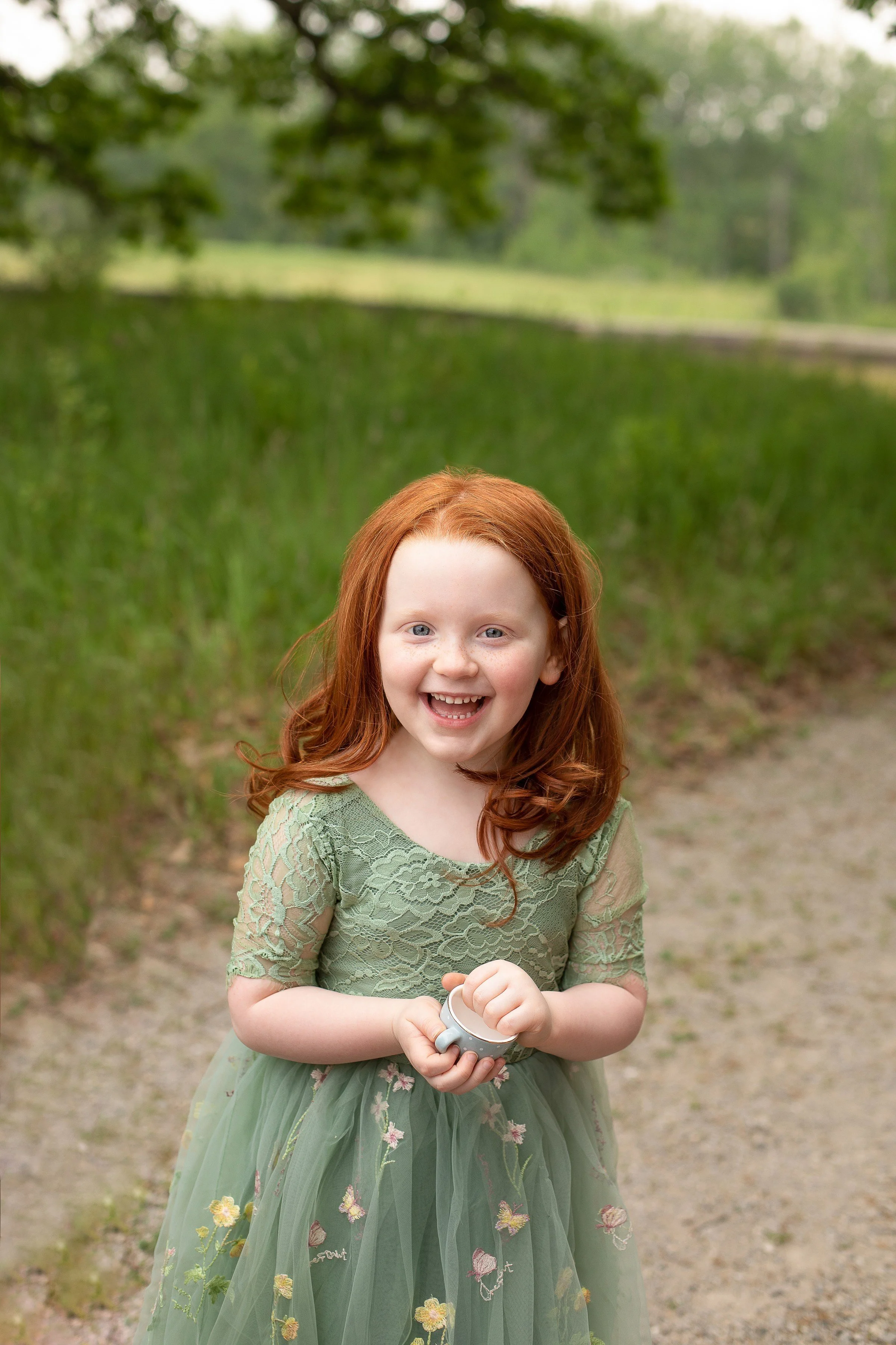 A smiling red-haired girl in a green lace dress standing outdoors near a body of water with green trees in the background.
