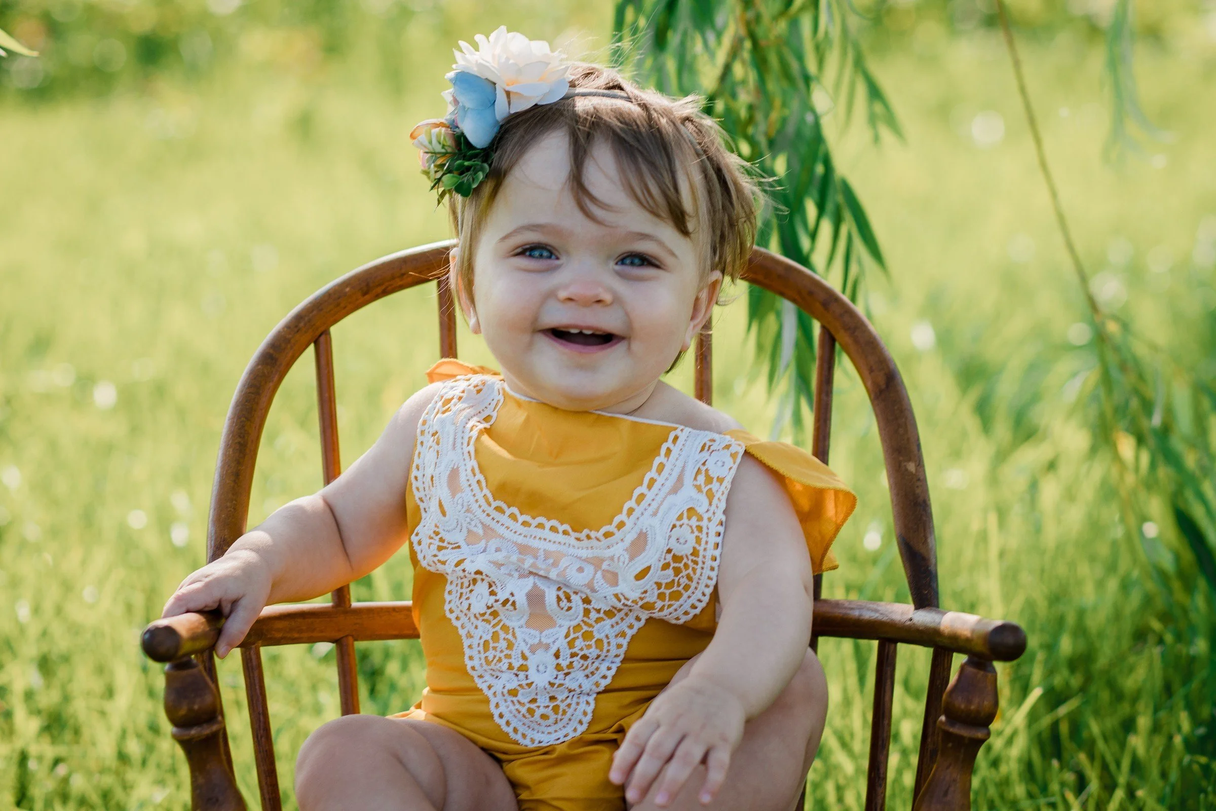 A smiling toddler girl with blue eyes and brown hair seated on a wooden chair outdoors in a grassy area, wearing a yellow dress with lace detail and a floral headband.