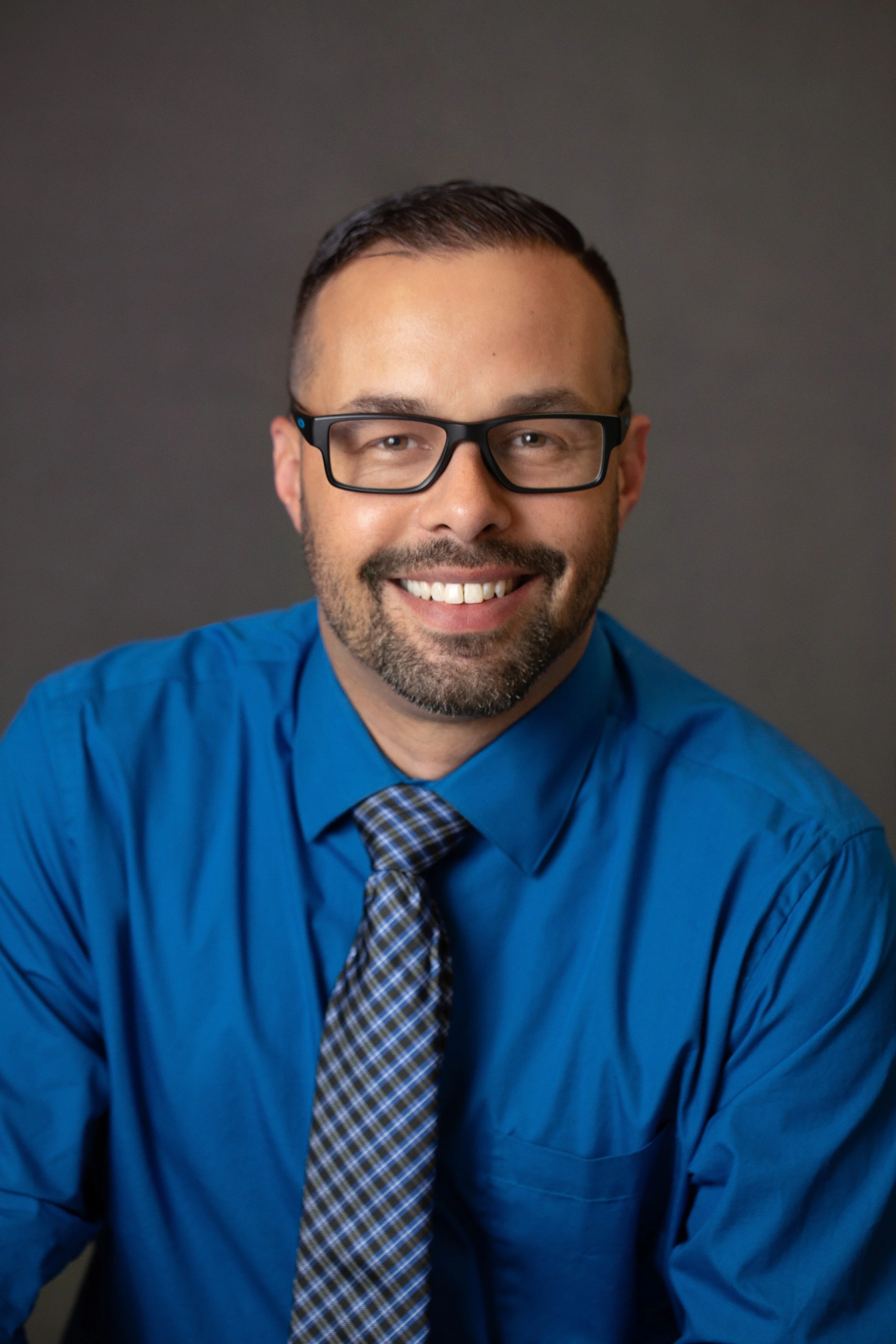 Portrait of a smiling man with glasses, wearing a blue shirt and patterned tie, against a dark plain background.