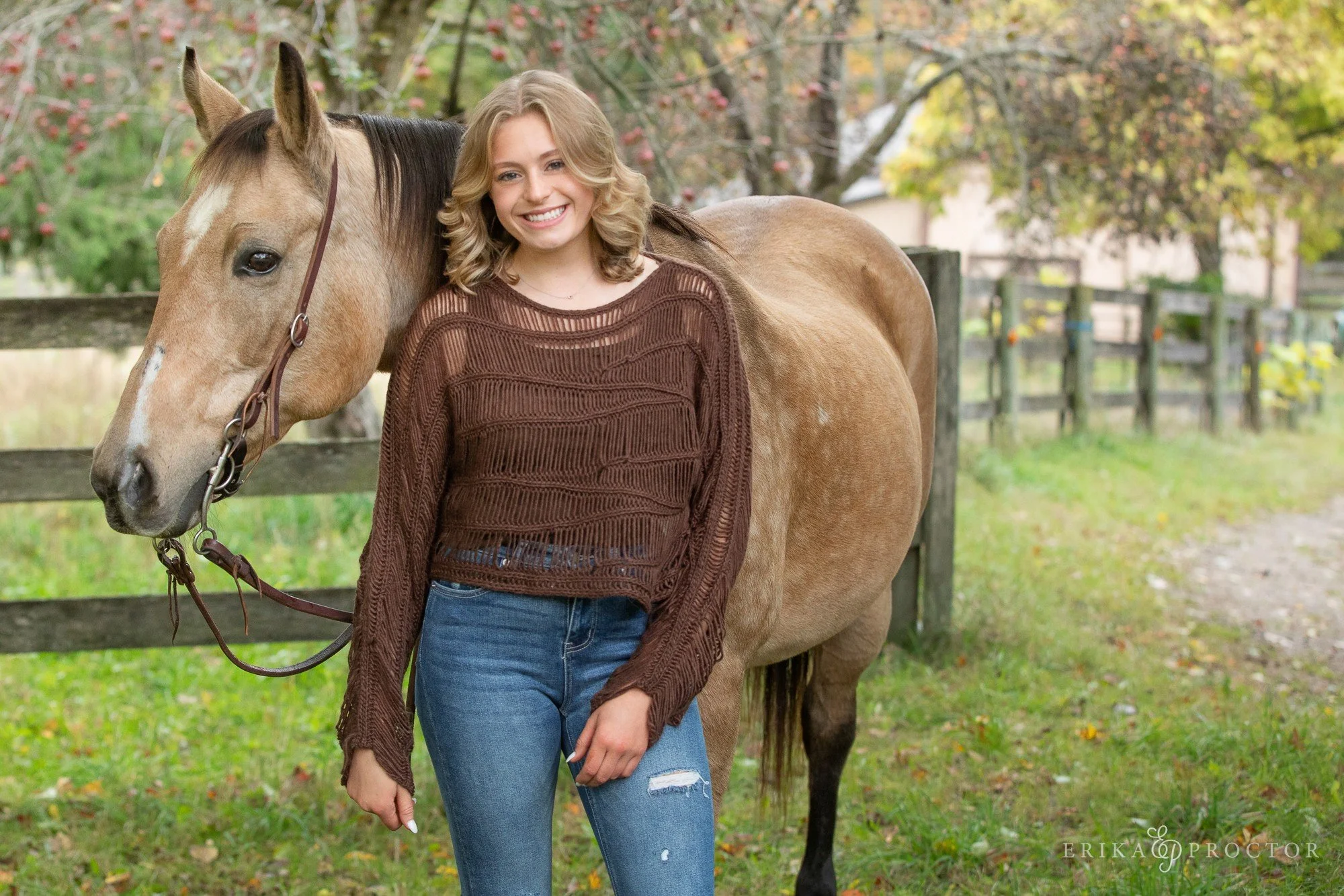A young woman with blonde curly hair smiling, standing next to a light brown horse with a dark mane, in a fenced outdoor area with trees and fall foliage.