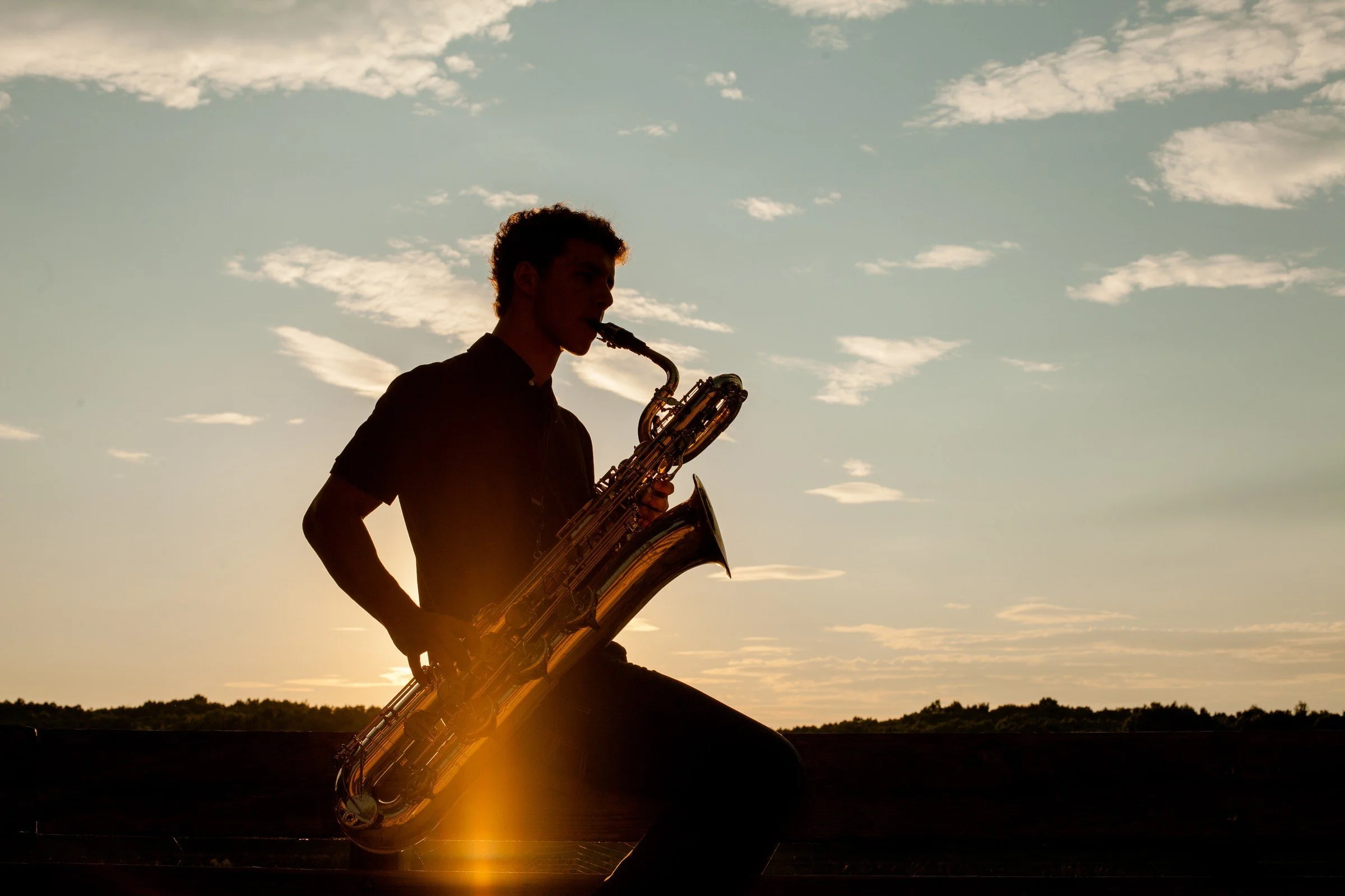 Silhouette of a young man playing the saxophone outdoors at sunset with a blue sky and scattered clouds in the background.